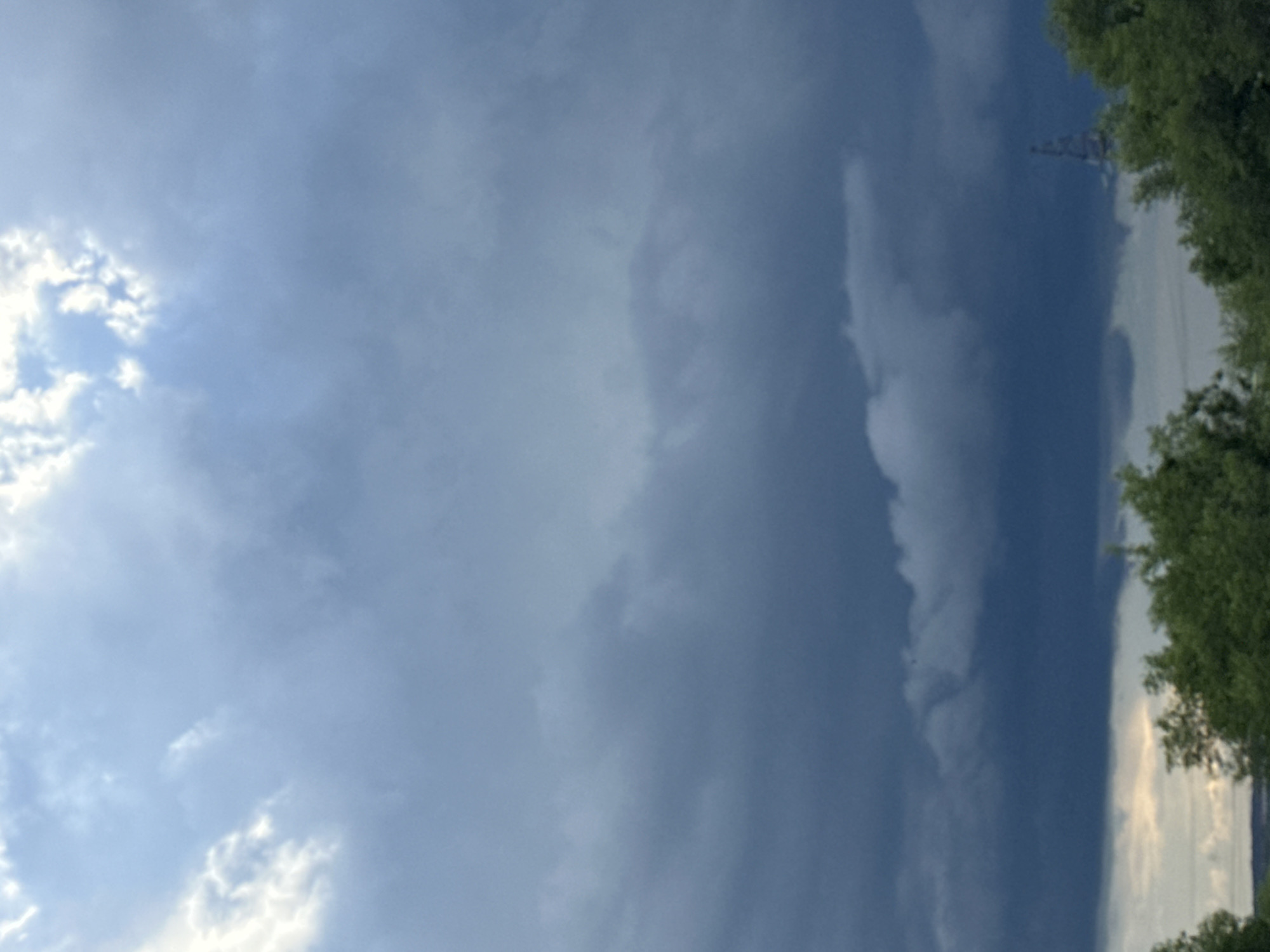 This media file is a vertically oriented photograph of a sky with clouds. The left side of the image shows bright, white clouds against a lighter blue sky, suggesting the sun might be near that area. As the view moves to the right, the sky darkens to a deep blue, with heavier, more ominous-looking clouds.
Partially visible on the far right is a line of green trees, indicating an outdoor setting, possibly a natural landscape or park. In the distance, towards the top right of the image, a tall, slender structure that appears to be a tower or pylon can be seen silhouetted against the sky. The overall impression is that of a dynamic sky, potentially with changing weather conditions, moving from fair to stormy. There is no visible text or human subjects in the image. The lighting suggests it could be daytime, though the dramatic cloud formations might imply either the late afternoon or the approach of a storm.