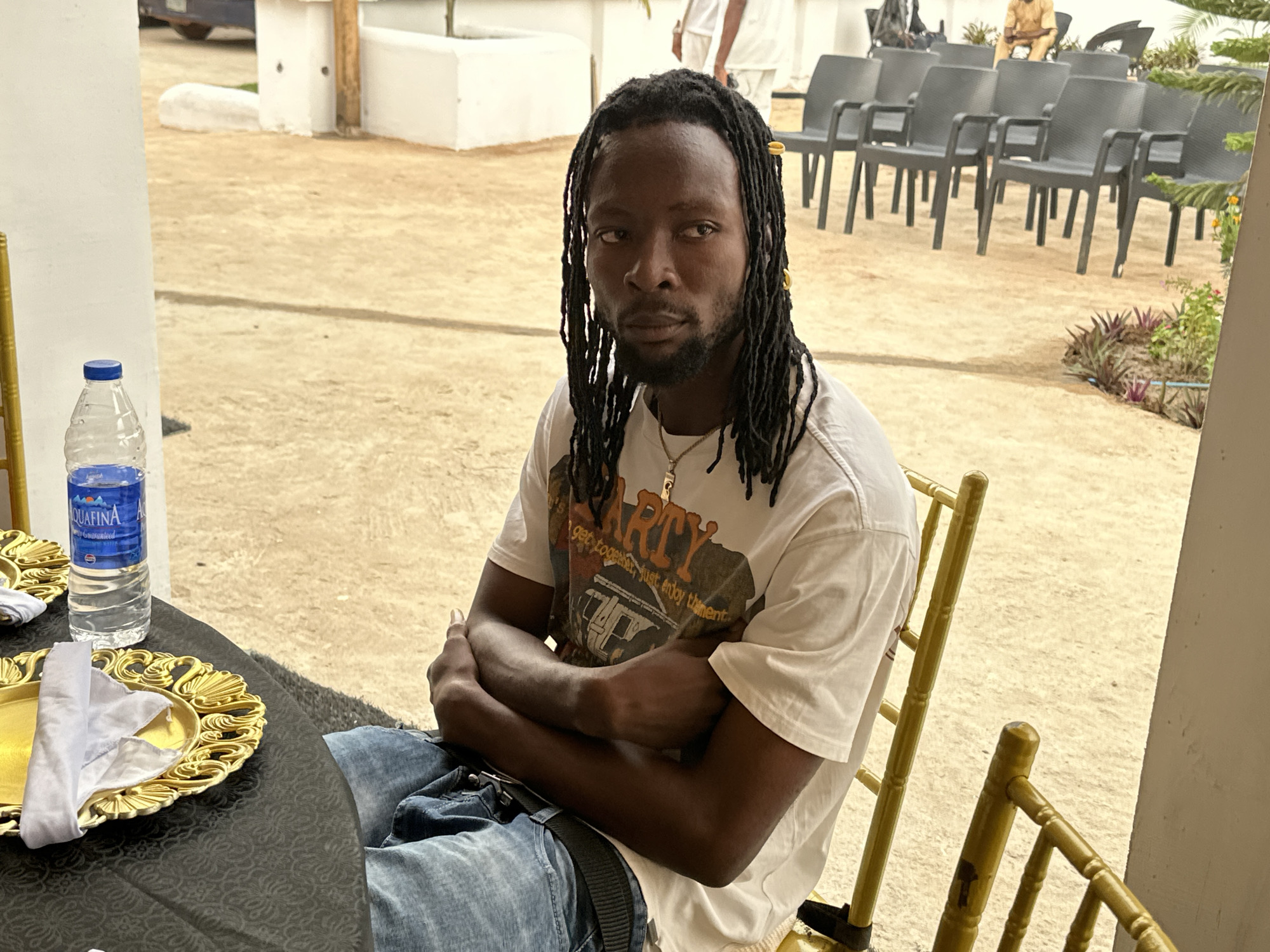 A man with dreadlocks, adorned with gold hair clips, sits with his arms crossed, looking directly at the camera. He is wearing a white t-shirt with graphic text that reads "PARTY Get together, just enjoy this moment," and blue jeans. He has a beard and is wearing a necklace.
In front of him, a black table is set with a golden decorative plate, a folded white napkin, and a bottle of Aquafina water. The man is seated on a golden chair.
The background reveals a sandy outdoor area with rows of black plastic chairs, suggesting a gathering or event. In the distance, people in white attire can be seen, along with some low-lying plants and white structures. The overall scene suggests an outdoor event or celebration taking place in Barden Yana, Nigeria, likely during daylight hours given the natural lighting.