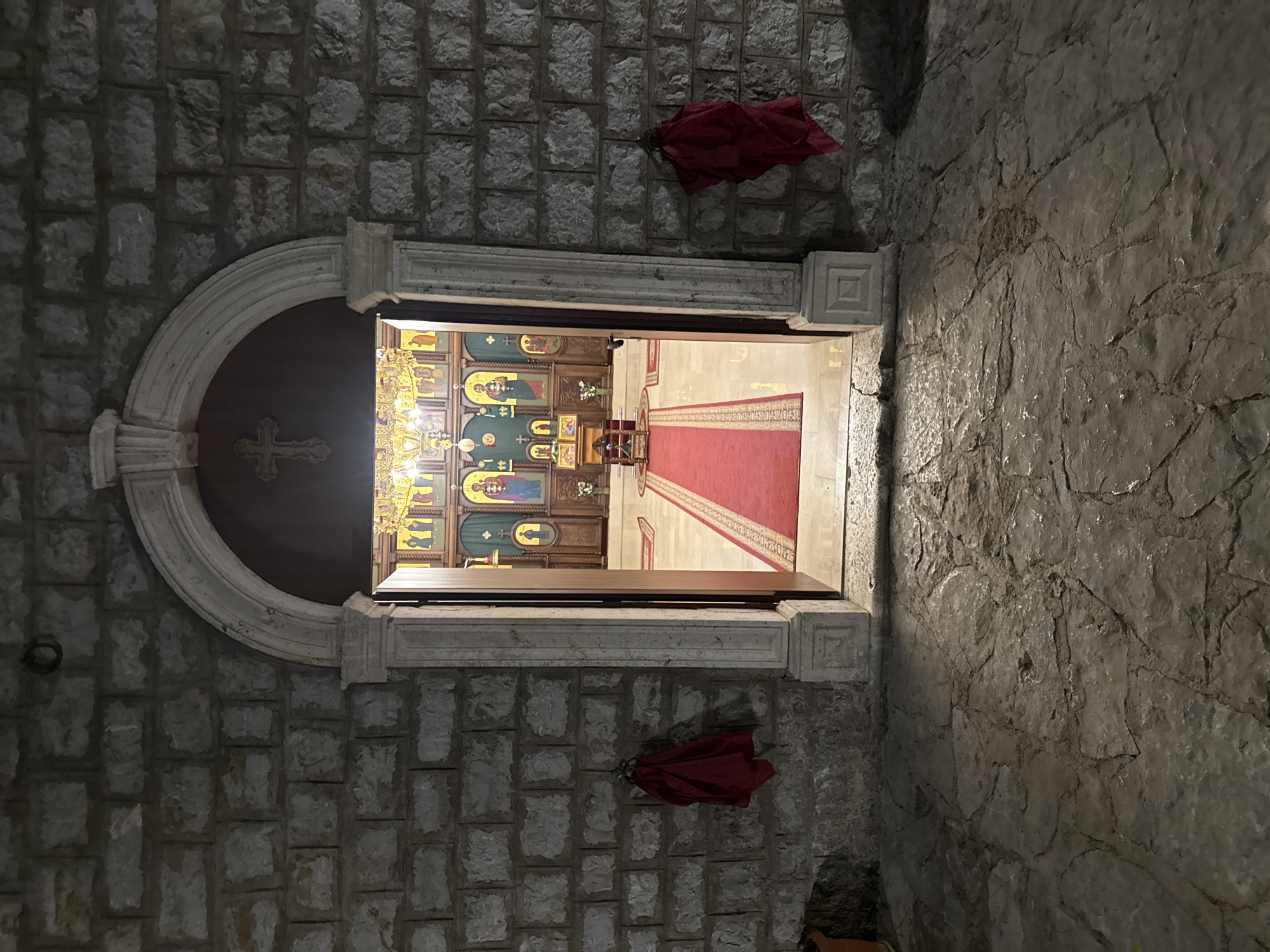 The image depicts the entrance to an Orthodox church, specifically identified as the Church of St. John the Baptist in Boreti, Montenegro. The exterior wall is constructed of rough-hewn grey stone blocks. An arched stone doorway, ornately carved and framed by a lighter stone, leads into the dimly lit interior. A red velvet carpet with a gold border runs from the doorway into the church. Inside, an iconostasis, adorned with religious icons and a prominent crucifix above, dominates the altar area. A large chandelier illuminates the scene. Two red cloths are draped on hooks on the exterior stone wall, one on each side of the entrance. The ground in front of the church is paved with irregular, weathered stone slabs. The overall impression is of an old, sacred building at dusk or night, with a sense of quiet reverence.