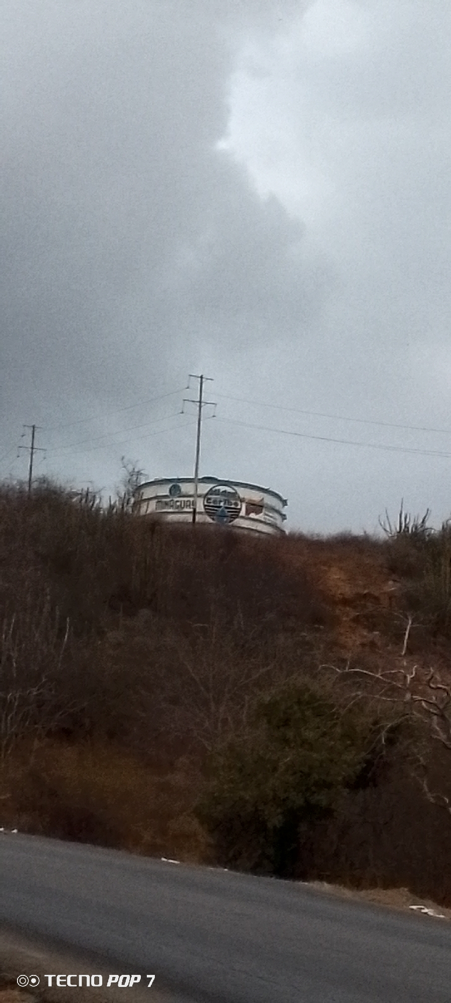 The image shows a water tank on a hill under a cloudy sky. The tank bears the inscription "AGUA Caribe." Power lines run parallel above the tank. The hillside is covered in scrub vegetation. A road is visible at the bottom of the frame. The photo was taken with a TECNO POP 7 phone. Based on the inscription "AGUA Caribe" and the style of the water tank, it is likely located in Altagracia, Venezuela.