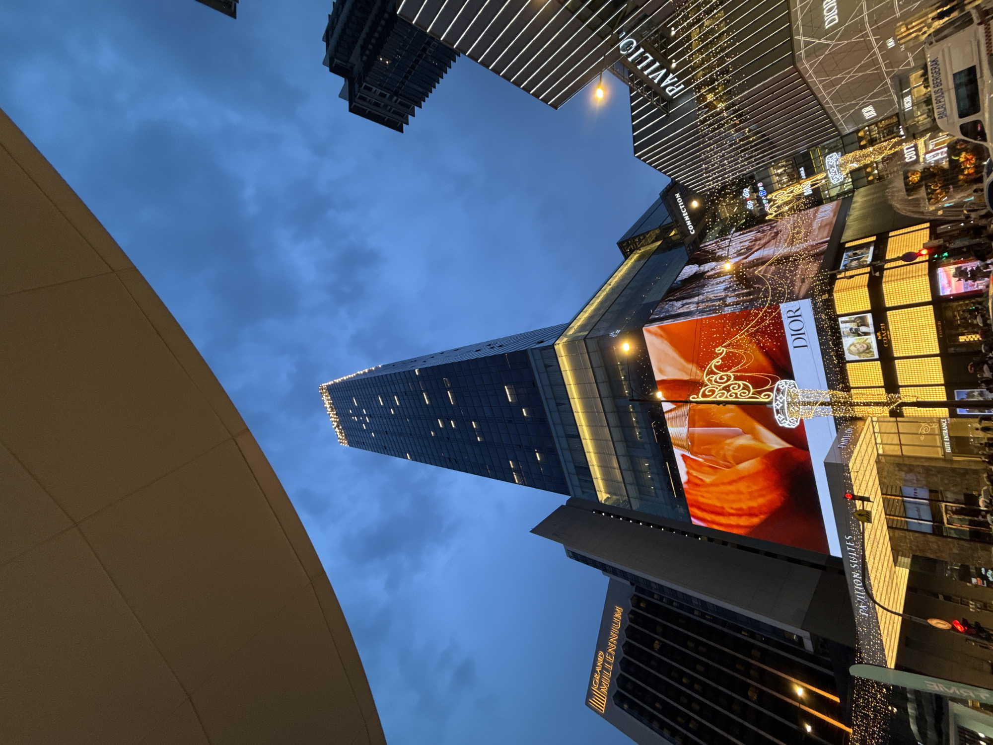 The image captures an upward angle view of Kuala Lumpur's urban landscape at twilight. Towering skyscrapers dominate the scene, including a slender, illuminated tower that extends into the cloudy blue sky. To the right, a building displays a large digital advertisement for Dior and is connected to the Pavilion, a prominent shopping center, marked by bright lights and visible store signs. A portion of the Grand Millennium Hotel is also visible. The bottom left corner features a part of a low, tan-colored structure, potentially a rooftop or canopy. The photo is taken at street level, as a mass of people can be seen walking. Overall, the scene depicts the modern architecture and commercial activity that characterize this area of Kuala Lumpur.