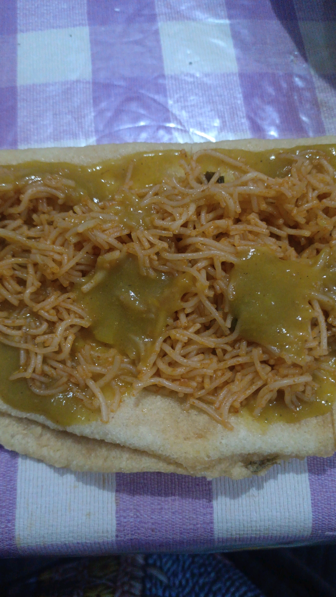 A close-up, overhead shot shows a savory dish, possibly a type of fried noodle or spaghetti, served on a light-colored, flat bread, resembling a thin roti or pancake. The noodles are fine and appear to be mixed with a reddish sauce and interspersed with dollops of a thick, yellowish-green sauce. The dish is placed on a surface covered with a checkered cloth that has alternating stripes of white and lavender. The fabric has a slightly crinkled texture, suggesting it might be plastic or coated. The overall lighting is dim, with a slight glare visible on the surface of the dish. The image is cropped tightly on the food, with the background of the checkered cloth and a hint of a dark surface beneath it. No people or other objects are visible.