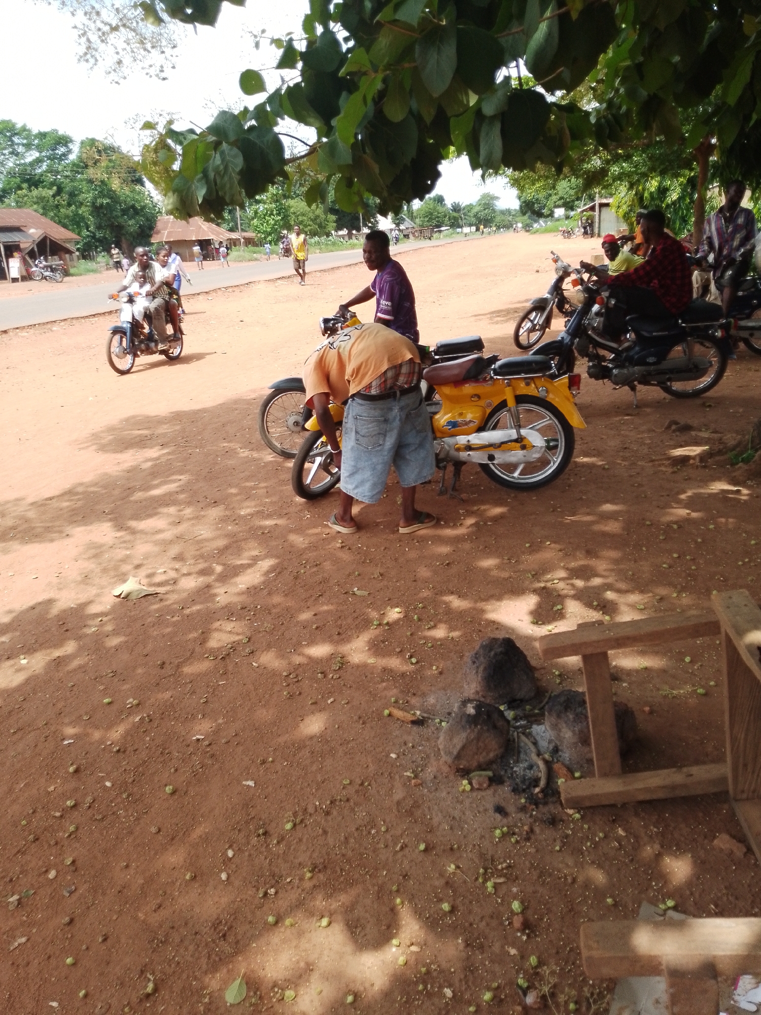 The image depicts an outdoor scene in Adikpo, Nigeria, likely during the day given the bright lighting and shadows. The ground is a dusty, reddish-brown earth, with scattered green seed pods and fallen leaves. Several people are present, many of them near motorcycles, suggesting a mode of transportation and perhaps a gathering place.
In the foreground, a person wearing an orange shirt and denim shorts is bending over a yellow motorcycle. This motorcycle appears to be a common model, possibly used for local transport. Behind this, another person in a purple shirt is visible. Further in the background, several other motorcycles are parked, and people are seen interacting or preparing to travel. One motorcycle has three individuals riding on it, including a child. In the far background, there are buildings with corrugated metal roofs, typical of rural or semi-urban areas in Nigeria. The presence of trees with large leaves suggests a warm climate, possibly in a tropical region.
The overall atmosphere appears casual and functional, hinting at daily life activities. There are no overt signs of celebration or distress, but rather a scene of people engaged in transportation or communal activities. The lighting suggests it could be late morning or early afternoon. The environment is natural and somewhat rustic.