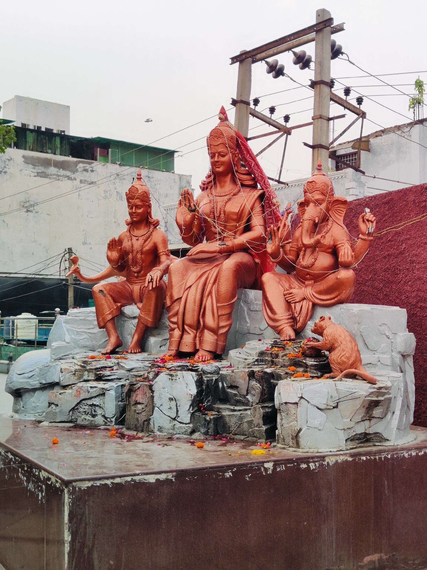 The image shows a religious statue installation depicting deities from Hinduism. A central female figure, presumably a goddess, is seated, adorned with a red veil and intricate gold-colored string work. To her left sits a male deity with multiple arms, holding a lotus-like object. To her right, a figure with an elephant head, identified as Ganesha, is seated with his hands in a gesture of blessing. In front of Ganesha, a small statue of a mouse, Ganesha's vahana (mount), is positioned, appearing to interact with offerings. The statues are made of a copper-colored material and are arranged on a rock-like structure with flowing water. The area is decorated with scattered marigold petals. In the background, buildings, electrical poles with wires, and a cloudy sky are visible. This scene is located in Gurgaon, India, specifically in the temple complex of Sheetla Mata Mandir. The overall impression is one of religious devotion and artistic representation of Hindu deities.