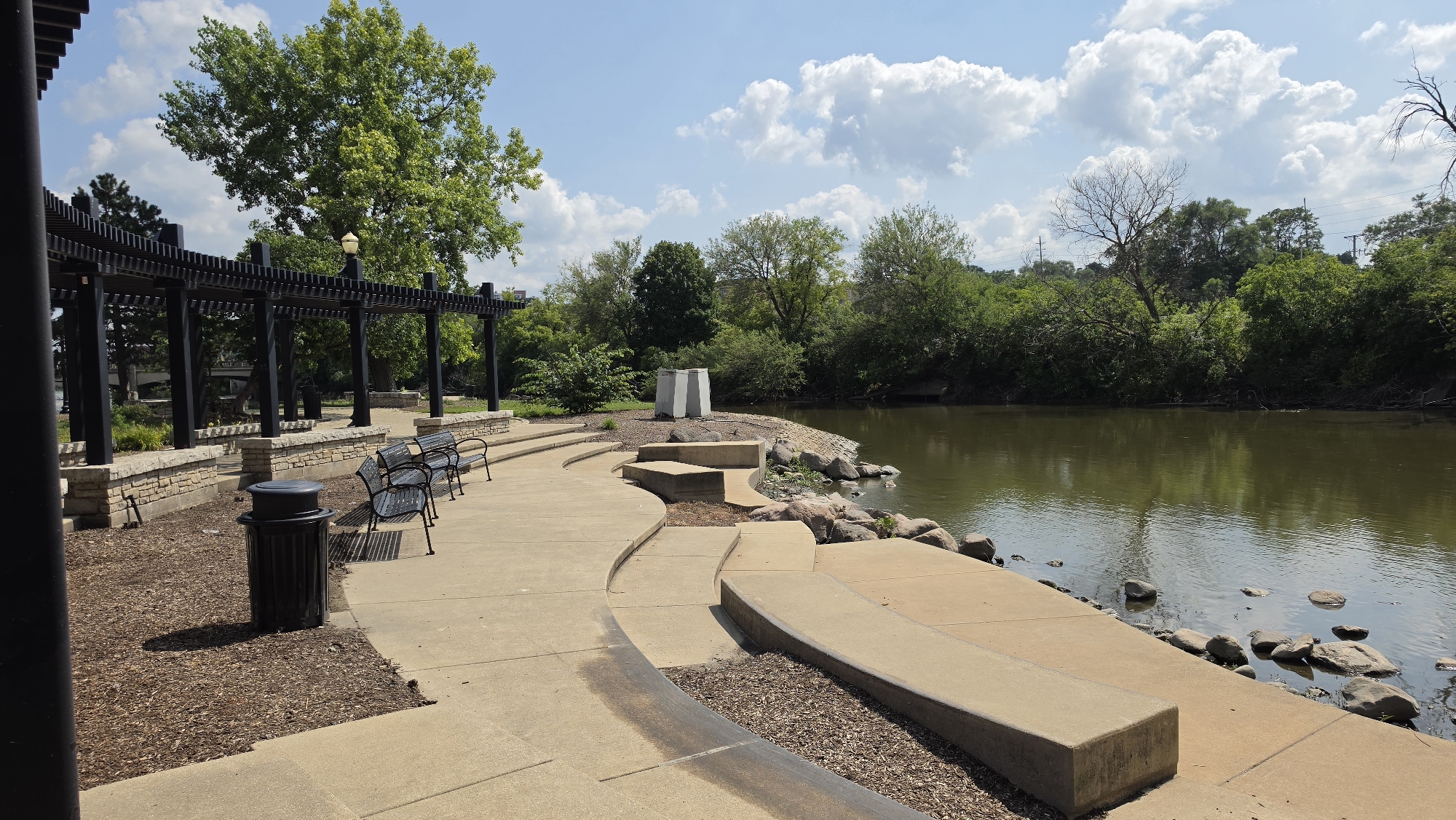This is a photograph of the Grand Victoria Casino Riverwalk in Elgin, Illinois, USA. The scene depicts a paved walkway with several benches and a trash can, leading to a tiered seating area overlooking a body of water. The water is calm and reflects the surrounding trees and sky. The sky is partly cloudy with large, white cumulus clouds. On the left side of the image, there is a black pergola structure with a light fixture. The ground is covered in wood chips in some areas and concrete in others. The riverbank is lined with rocks and vegetation. In the distance, trees and foliage line the opposite bank of the river. The photograph appears to have been taken on a sunny day.