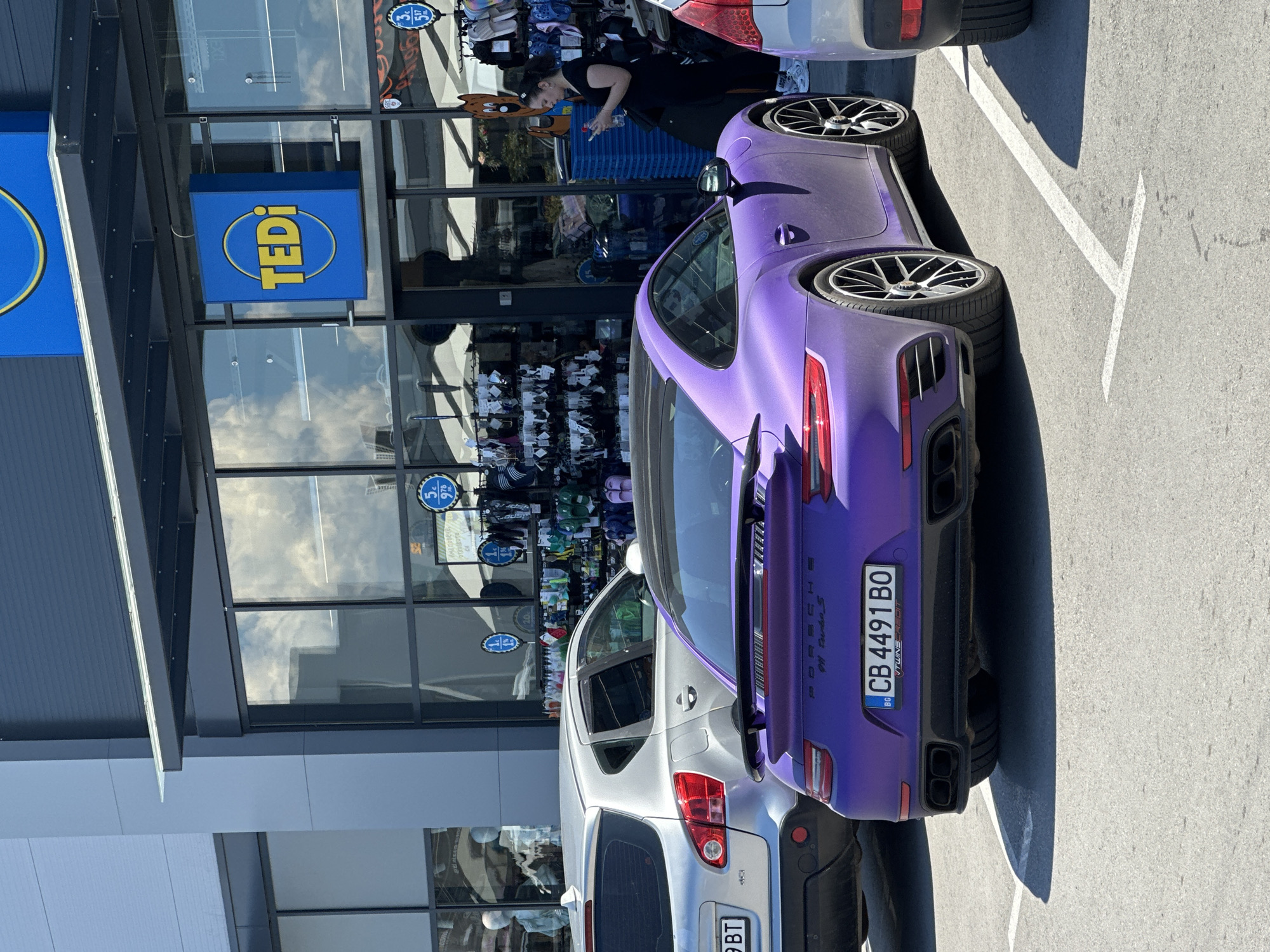 A matte purple Porsche 911 Turbo S is parked in a lot in front of a Tedi store, located in the city of Veliko Tarnovo, Bulgaria. The car's license plate reads "CB 4491 BO". To the left of the Porsche is a silver Nissan Qashqai with a license plate that partially reads "9 BT". To the right of the Porsche, a portion of a silver sedan is visible. A woman wearing dark clothing and white sneakers stands near the entrance of the Tedi store, holding a phone and a water bottle. The Tedi store has large glass windows displaying various merchandise, and the store's blue and yellow logo is prominently displayed. The scene appears to be outdoors on a sunny day, with clouds visible in the reflection of the store windows.