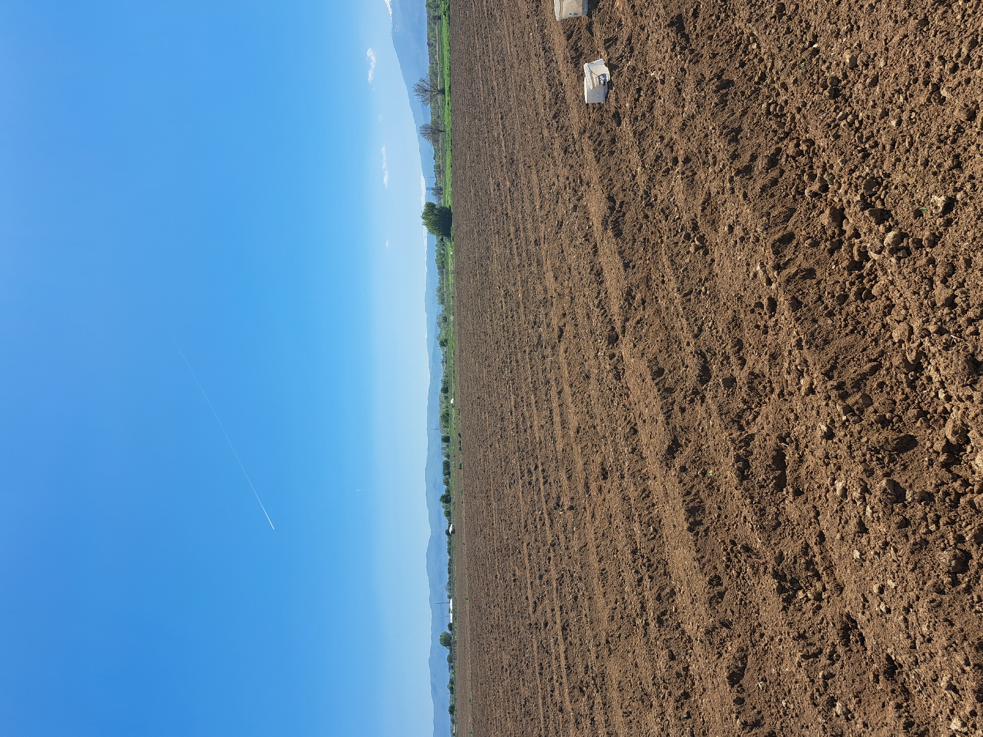 The image shows a recently plowed field under a clear blue sky. In the distance, a treeline and mountains are visible. There are two white objects, possibly containers, in the plowed field. A contrail is visible in the upper left portion of the sky. The location is Unknown, Unknown.