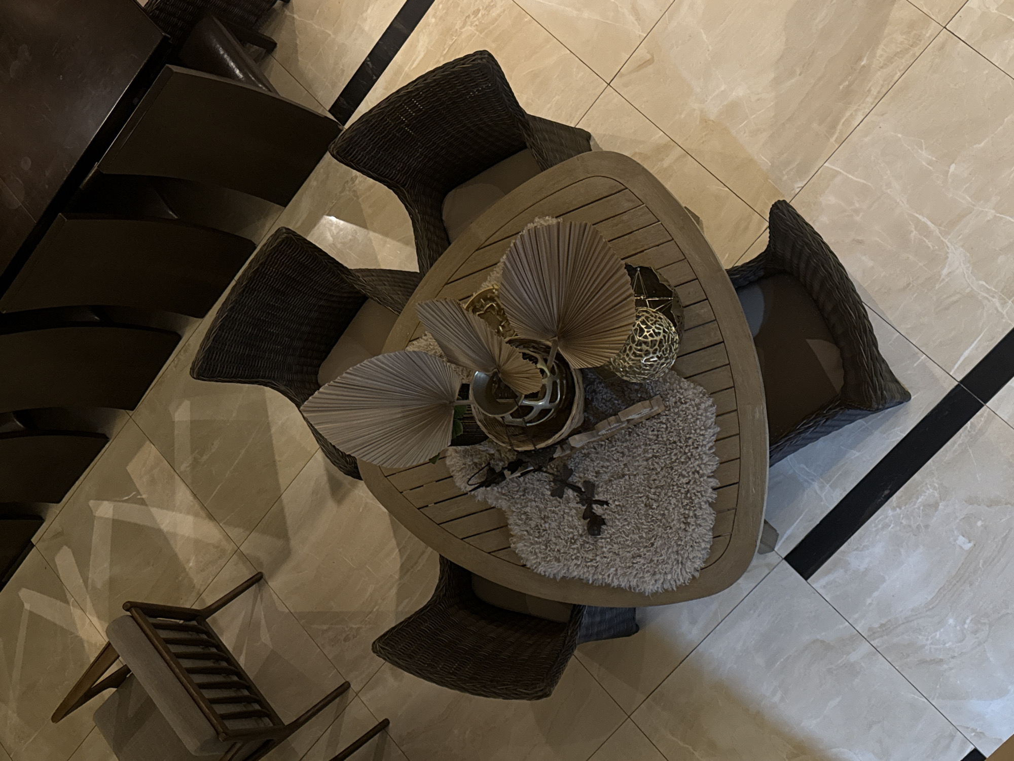 The image depicts an overhead view of a round wooden table with several chairs surrounding it, likely in a dining or living area. The table is adorned with decorative items, including a fuzzy rug, dried fan palm leaves, and metallic spherical ornaments. The chairs appear to be made of woven material, and one wooden armchair with a grey cushion is visible to the left. The flooring consists of large, light-colored tiles with dark grout lines, and there are hints of sunlight creating geometric patterns on the floor. The scene is set indoors, with the overall impression being one of a modern and tastefully decorated space. There is no indication of the specific location in Bac Nha Trang Ward, Vietnam.