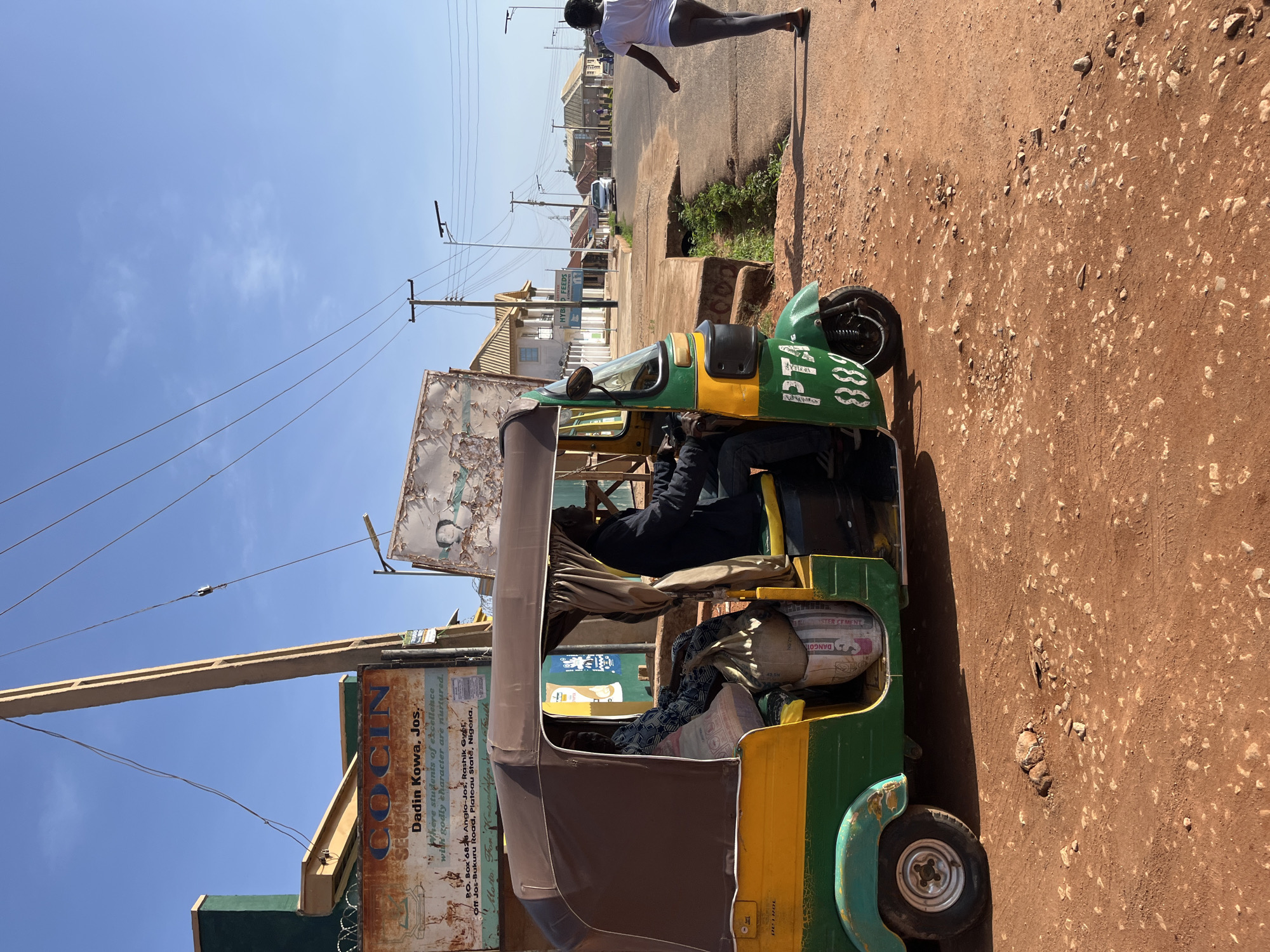 The image presents a sun-drenched street scene in what appears to be a residential or commercial area, consistent with the provided context of Jos, Nigeria. The dominant subject is a green and yellow three-wheeled auto-rickshaw (often called a keke napep or tuk-tuk), which is either parked or moving slowly on the side of a reddish-brown, unpaved road.
Inside the auto-rickshaw, a person in dark clothing occupies the driver's seat. The vehicle is also loaded with what appears to be cargo, including sacks prominently labeled "DANGOTE LUSTER CEMENT." On the front right side of the vehicle, the letters "PTA" and the number "088" are visible.
Further down the street, on a raised sidewalk or curb, a person dressed in a white t-shirt and dark leggings is walking. The road surface is dry and dusty, scattered with small stones and debris. Buildings line both sides of the street in the background, with power lines and utility poles crisscrossing the clear, bright blue sky.
A large, weathered sign on the left side of the frame features prominent text. It reads: "COCIN SECOND," followed by "Dadin Kowa, Jos," and a description: "Where students of excellence with godly character are nurtured." It also includes an address: "P.O. Box 6828 Anglo-Jos, Rashik Gy, Off Jos-Bukuru Road, Plateau State, Nigeria." A motto, "Motto: For," is partially visible. Another sign further back, on a building, displays "HYBOD FEEDS."
The bright sunlight and clear sky indicate it is daytime with good weather. The overall scene depicts a typical street environment in a bustling locality, with everyday transportation and commercial activities taking place.