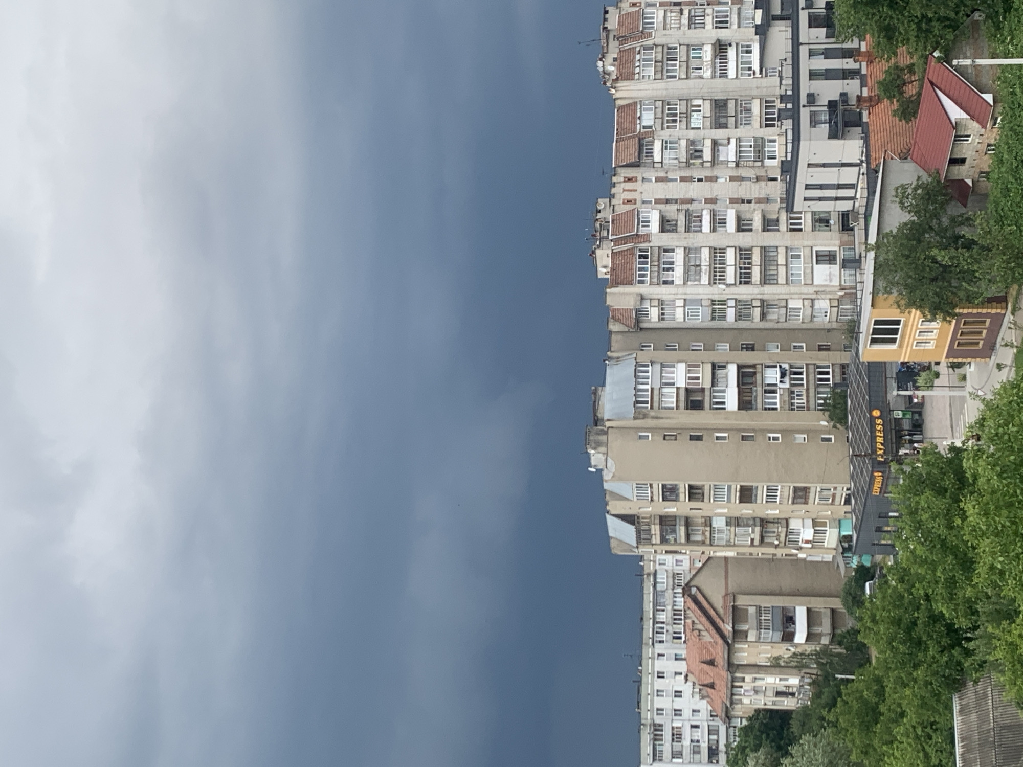 This is an eye-level, wide-angle exterior shot of an urban residential area in Kolomyia, Ukraine, under a dark, overcast sky. The buildings are primarily Soviet-era apartment blocks, characterized by their repetitive facade designs, balconies, and light-colored exteriors. In the foreground, lush green trees dominate the lower portion of the frame. A small, modern-looking gas station with "EXPRESS" signage is visible nestled among the trees, with people and vehicles around it. In the mid-ground, several apartment buildings of varying heights are lined up, their windows reflecting the dim light. The largest building in the center has a beige facade and numerous balconies. The sky above is a deep, ominous grey, with heavy clouds suggesting impending rain or a storm. The overall impression is one of a typical Eastern European city district, with a mix of older and newer construction, and a natural element in the foreground.