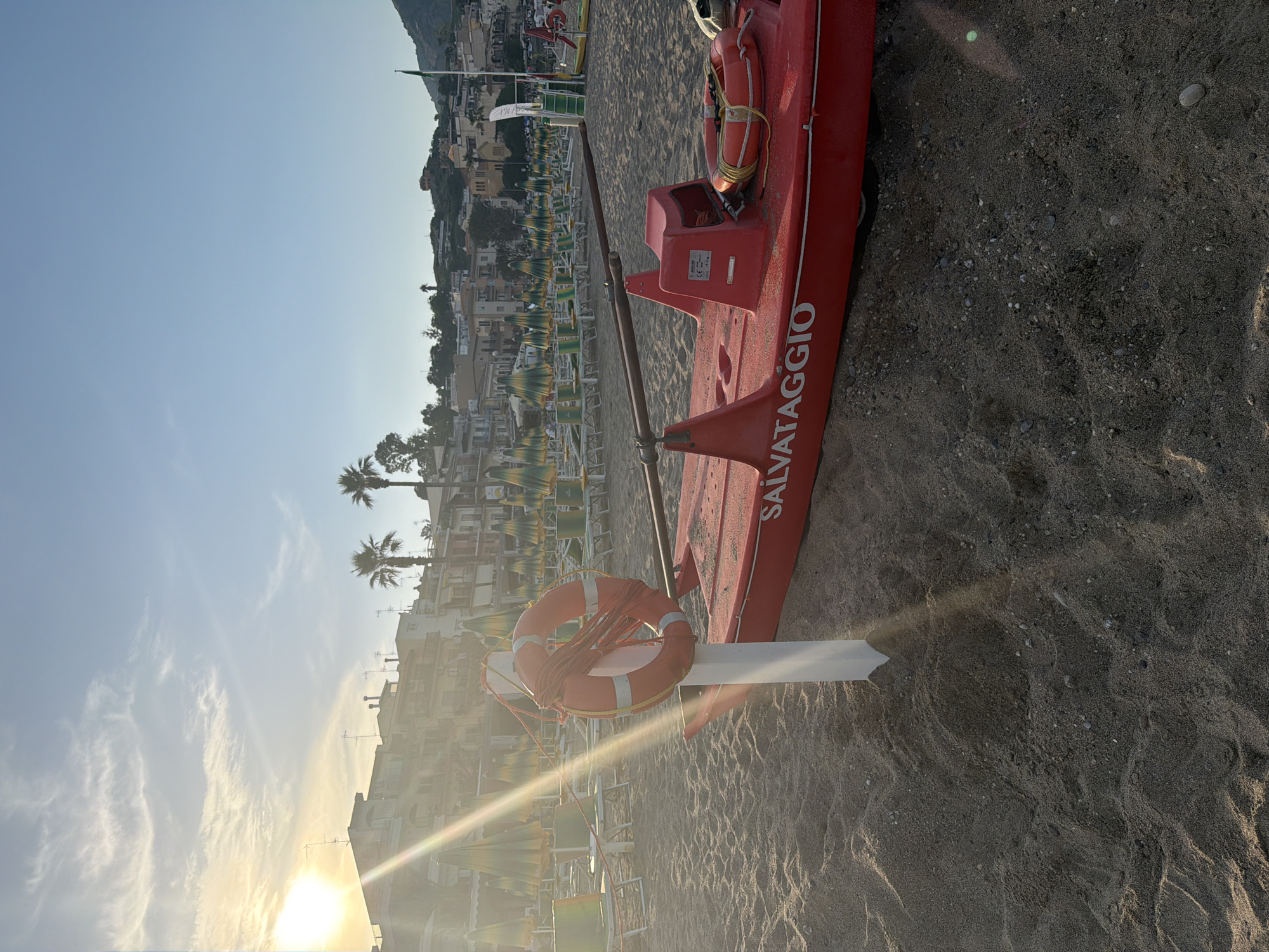 The scene is a beach in Chianchitta, Italy. A red lifeguard boat, labeled "SALVATAGGIO," rests on the sand, with a life preserver affixed to it. Beach umbrellas and chairs are visible in the background, near buildings and palm trees. The sky is blue with some clouds, and the sun is low, creating lens flare. The sand is dark and textured.