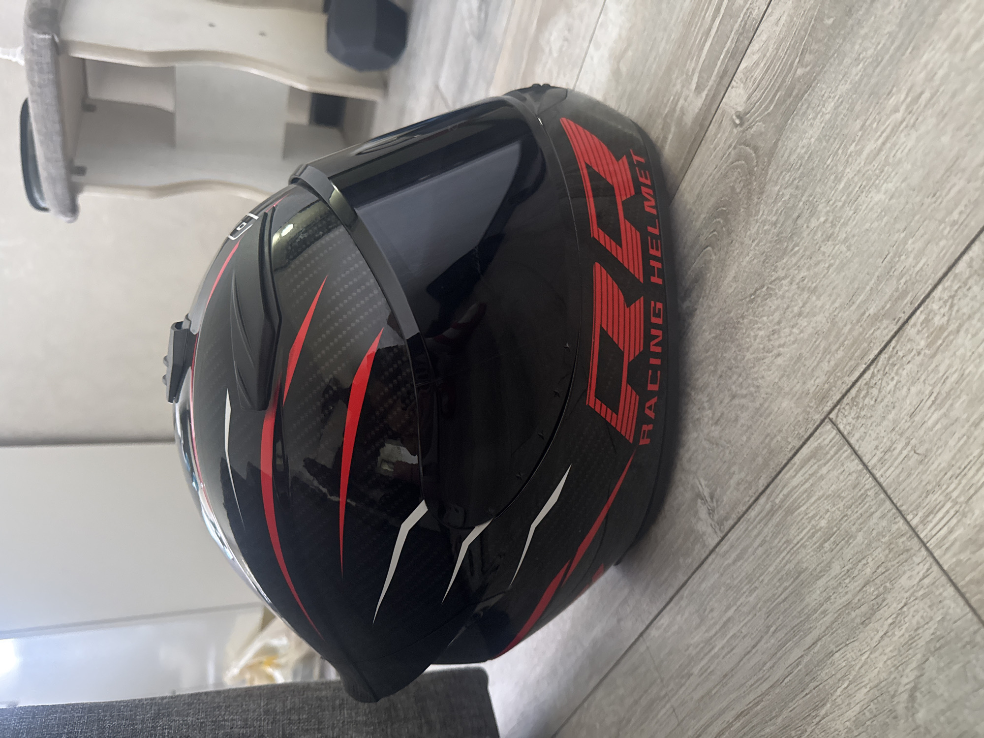 A black and red motorcycle helmet is on a gray wooden floor. The helmet has a carbon fiber pattern and red and white accents. Text on the helmet reads "RR RACING HELMET". The background is blurred and shows a white wall and furniture. The lighting is bright and comes from the left.
