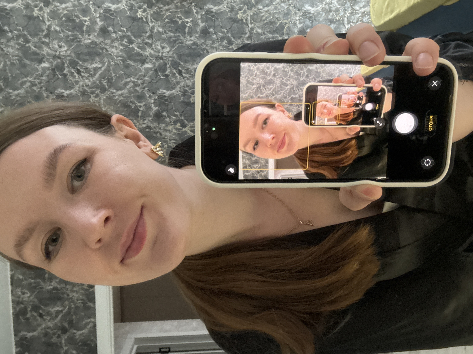 A woman is holding a phone, taking a picture of herself taking a picture of herself. The woman has light skin, brown hair, and is wearing a gold bee earring. She is wearing a necklace. The phone is black with a white case. The background appears to be a bathroom with a gray marble countertop. The location in Izhevsk, Russia is not discernible.