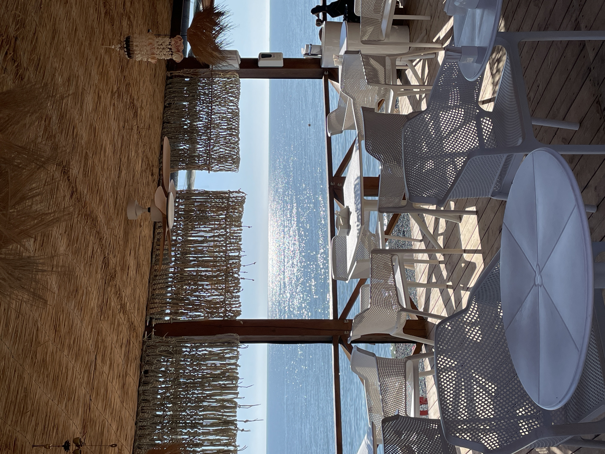 This image captures a detail-rich scene of an unoccupied outdoor cafe or restaurant directly overlooking the Black Sea in Sochi, Russia. The establishment features a sturdy roof constructed from tightly woven, light-brown straw or thatch. This roof is supported by dark-stained wooden posts and beams. Hanging beneath the roofline are decorative, light-colored, macrame-style screens or curtains with a fringed appearance, partially framing the view of the sea.
The floor of the seating area is composed of light-brown wooden decking. The furniture consists entirely of white, modern-design pieces: numerous plastic chairs featuring a distinct mesh or lattice pattern, and a combination of round and square tables. Ashtrays are visible on at least one table, alongside small white rectangular containers, likely for napkins or utensils.
Ceiling fixtures include a white, three-bladed ceiling fan with light-colored wooden-textured blades on the right side. Adjacent to it, a decorative mobile made of white and pink seashells with dark accents hangs. A smaller, darker metallic bell decoration is suspended from the roof structure on the far left.
The background reveals a vast expanse of calm, deep blue sea under a clear sky. A prominent path of sunlight reflects brightly off the water's surface, creating a sparkling effect that extends towards the distant horizon. A dark wooden railing separates the cafe's deck from the narrow, pebble-strewn shoreline below. A small, spherical orange buoy is visible floating in the mid-distance on the sea.
Mounted on one of the wooden support posts on the right is a white wall-mounted dispenser, likely for hand sanitizer. Below this, a small white basin with a chrome faucet, suggesting a compact hand-washing station, is present. Adjacent to the basin stands a dark, bird-shaped figurine, possibly a duck. The scene is devoid of people, indicating the tables are currently unoccupied.