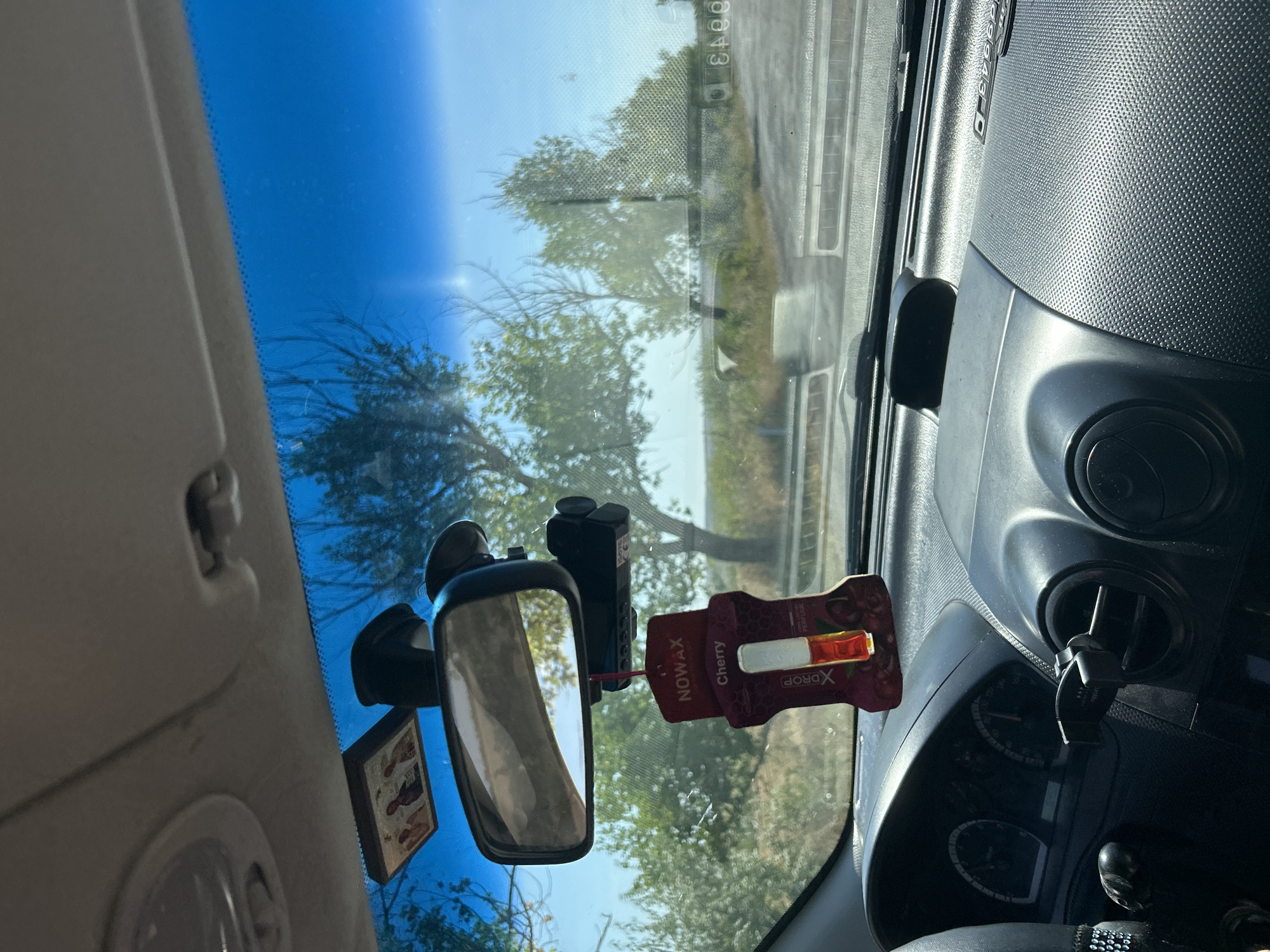 An interior view from a car dashboard shows a rearview mirror with a dashcam attached. Hanging from the mirror is a "NOWAX XDROP Cherry Perfume" air freshener. In the background, through the windshield, a road and trees are visible under a clear blue sky. A religious icon is affixed to the ceiling of the car above the rearview mirror.