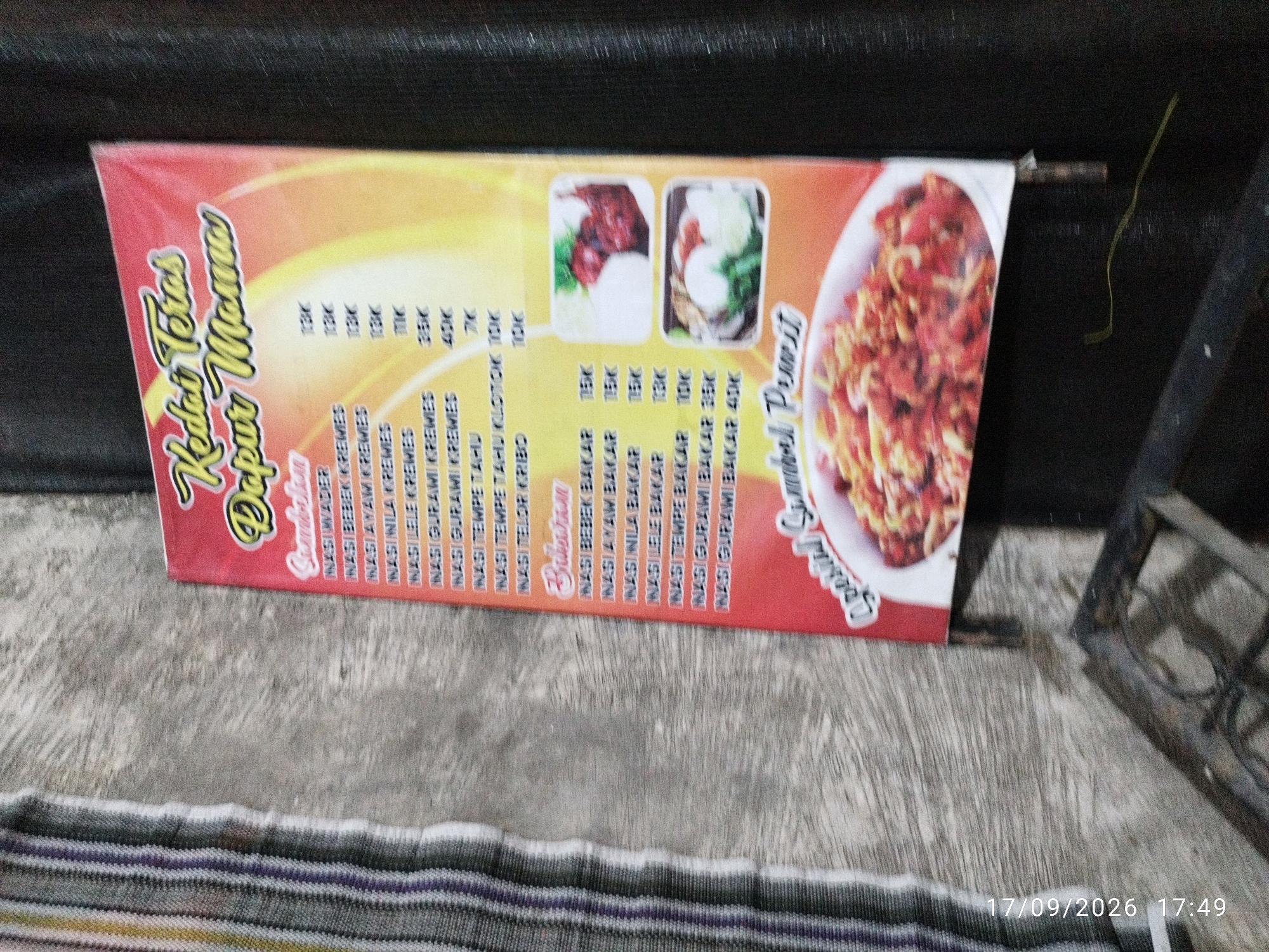 A blurry horizontal banner is displayed outdoors, mounted on a dark, textured surface. The banner, with a red, orange, and yellow gradient background, features text in Indonesian and images of food. Text on the left side reads "Kedai Teras Dapur Mama," suggesting it is a menu from a food stall named "Mama's Kitchen Terrace." Below this title is a list of food items with prices. The central part of the banner shows two small rectangular images of dishes, likely menu items. To the right, a larger image displays a bowl of what appears to be fried noodles or a similar dish. Below the images, text continues to list food items and prices, including "Nasi Goreng," "Ayam Bakar," and "Lele Goreng." The banner is attached to a dark metal bar on the right side. The background appears to be a rough, grey concrete surface, and a dark, textured wall or fabric is visible behind the banner. A thin, yellow string or wire hangs from the metal bar. The bottom of the image shows a portion of a striped fabric. A timestamp in the lower right corner reads "17/09/2026 17:49." The location is identified as Mojokerto, Indonesia, based on the context provided.