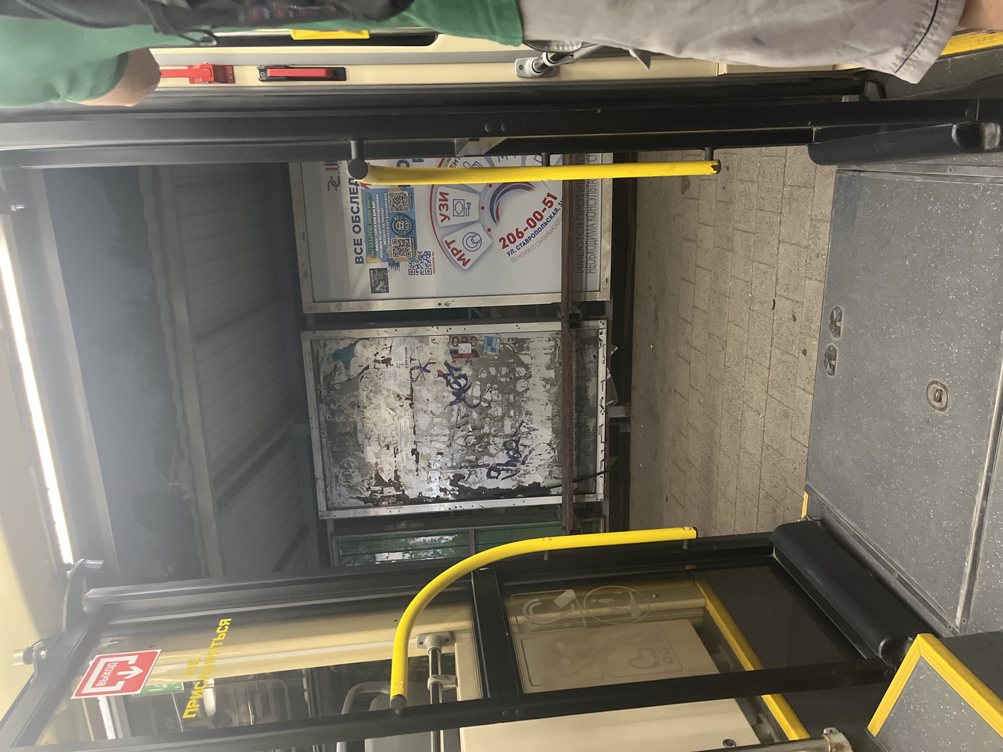 The image shows an open doorway of a bus or tram, facing a bus stop shelter. A red sign with a white arrow pointing down and the Russian word "ВЫХОД" (EXIT) is visible on the left door frame. Below it, yellow text reads "НЕ ПРИСЯЖАТЬСЯ" (DO NOT LEAN). Inside the bus, yellow handrails are present.
Outside the bus, a weathered advertising poster is attached to the back wall of the shelter. To the right of this poster is a modern advertisement with the text "ВСЕ ОБСЛЕД" (ALL EXAMINATIONS) and a phone number "206-00-51" along with an address "ул. СТАВРОПОЛЬСКАЯ, 3". QR codes are also present on this advertisement.
The bus stop pavement is made of light-colored bricks. The lighting suggests it is either overcast or late in the day. The presence of Cyrillic text on the advertisements and signs indicates a location in a Russian-speaking country. Specifically, the street name "Ставропольская" is a common street in Russian cities, and Krasnodar is a major city in Southern Russia known to have a "Ставропольская улица". Therefore, the scene is likely a bus stop in Krasnodar, Russia. A person wearing a green shirt and light-colored shorts is partially visible on the right side of the frame, suggesting they are entering or exiting the vehicle.