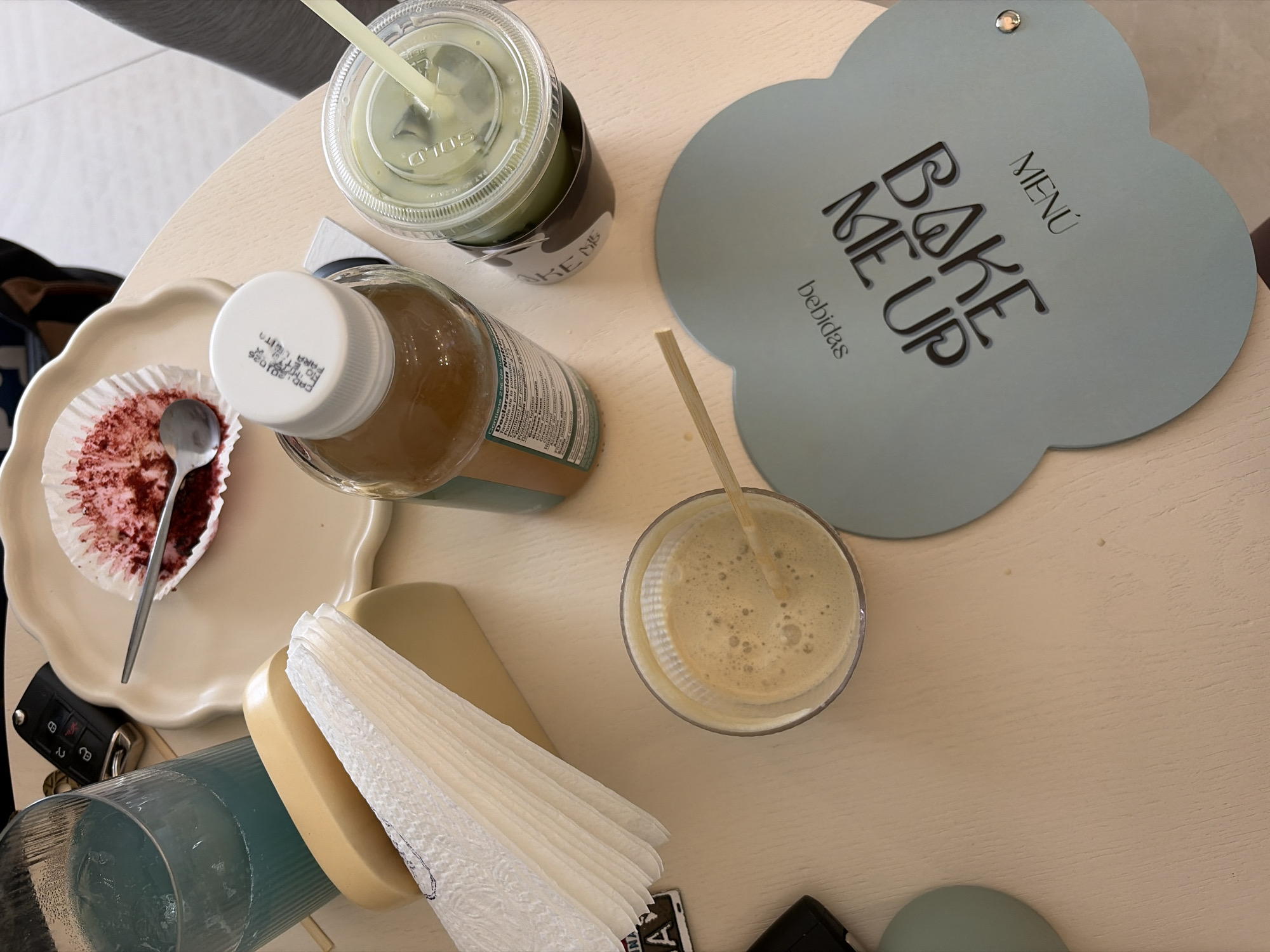 An overhead shot captures a light-colored wooden table laden with refreshments and personal items, likely in a cafe or bakery. In the upper right, a light blue, cloud-shaped menu displays "MENÚ BAKE ME UP bebidas" (drinks) in black lettering.
Several beverages are present: a clear plastic cup holds a green drink (possibly matcha) with a straw, its sleeve partially showing "BAKE ME". A glass bottle with a white cap contains a brown liquid, and its label, though mostly obscured, shows nutritional information including "Contiene 296 de ju" (likely juice), "Tamaño de la porción", "Grasa", and "PARA LEGITA" on the cap. Another glass contains a frothy, light brown beverage with a straw.
On a white, scalloped plate, a red velvet cupcake wrapper holds remnants of the cake and a small spoon, suggesting it has been mostly consumed. A stack of white paper napkins rests on a beige holder. A black car key fob is visible near the plate, and a blue-tinted glass is partially seen in the lower left. Part of a person's leg or clothing is visible in the top left corner, indicating someone is seated at the table enjoying the items. The scene suggests a casual cafe visit during the daytime.