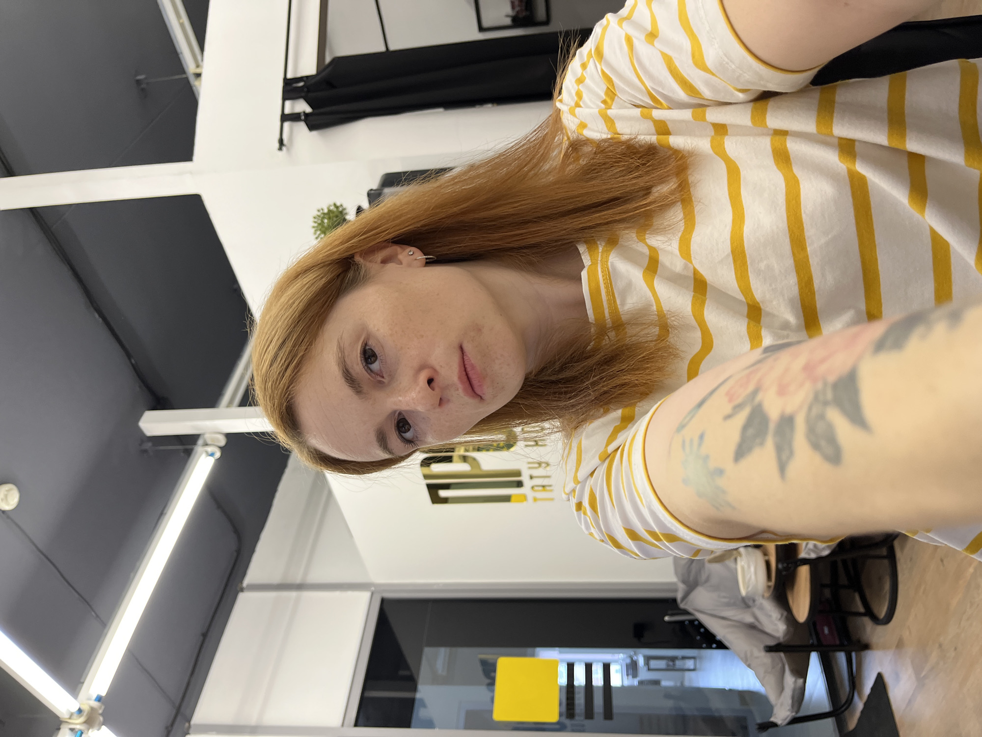 A selfie portrait of a fair-skinned individual with light brown hair, wearing a white and yellow striped shirt. The person's left arm is visible, featuring a tattoo of floral designs. The background reveals an interior space, identifiable by "TATY HOU," indicating a possible place of business or tattoo parlor. Through a glass door, one can see part of a room with a yellow square on it. The location is likely in the city of Chaikovsky, Russia, based on the business name. The image is taken from an angled perspective, slightly tilted to the side.