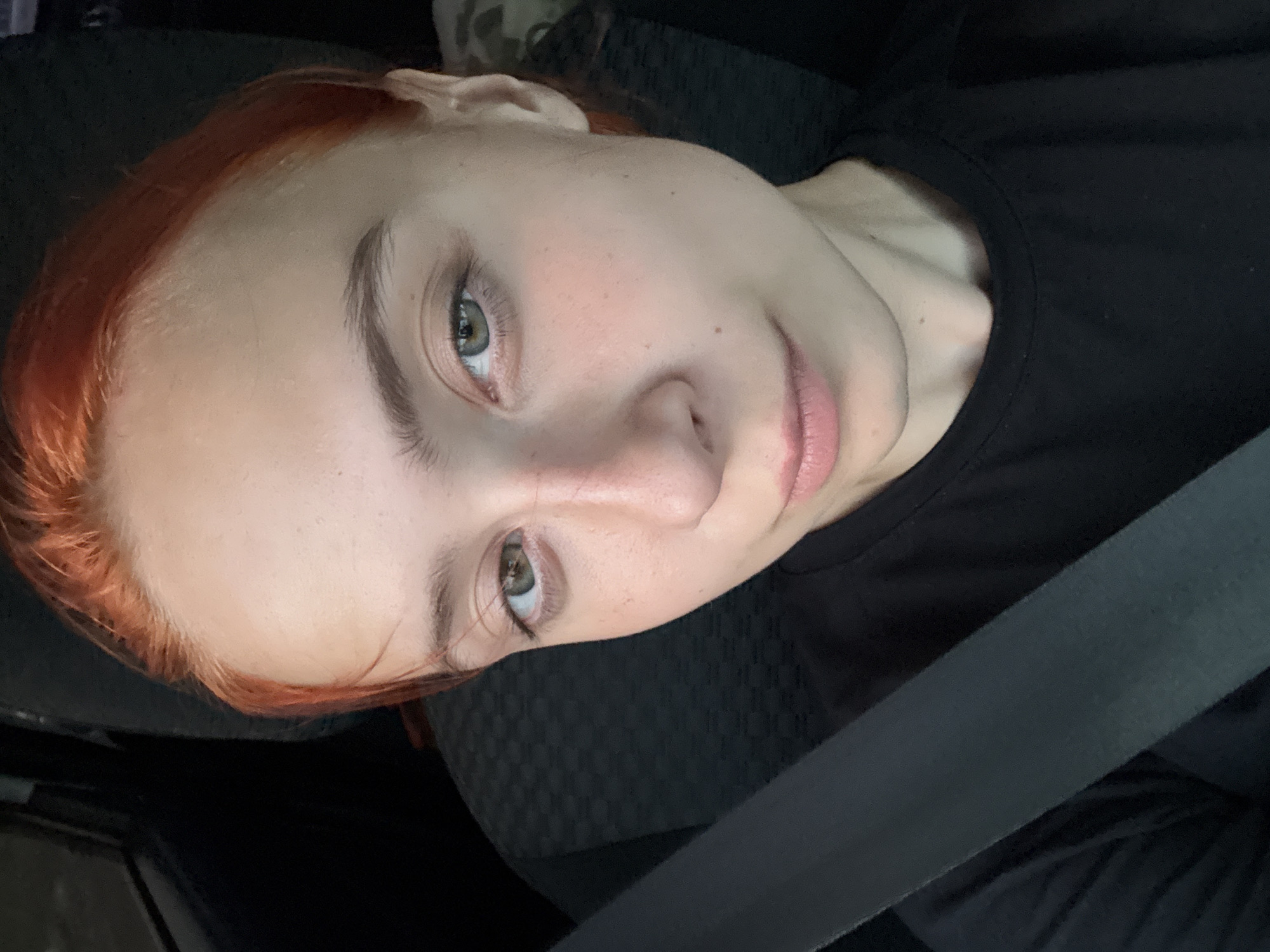 A young woman with reddish-orange hair and light skin with freckles is pictured inside a vehicle, possibly a car, as indicated by the presence of a seatbelt across her chest. She is wearing a black t-shirt. Her gaze is directed towards the camera. The background appears to be the interior of a vehicle, with a dark, textured seat behind her. There are no discernible landmarks or features that would place this image specifically within Votkinsk, Russia, or any other city. The lighting suggests it is daytime.