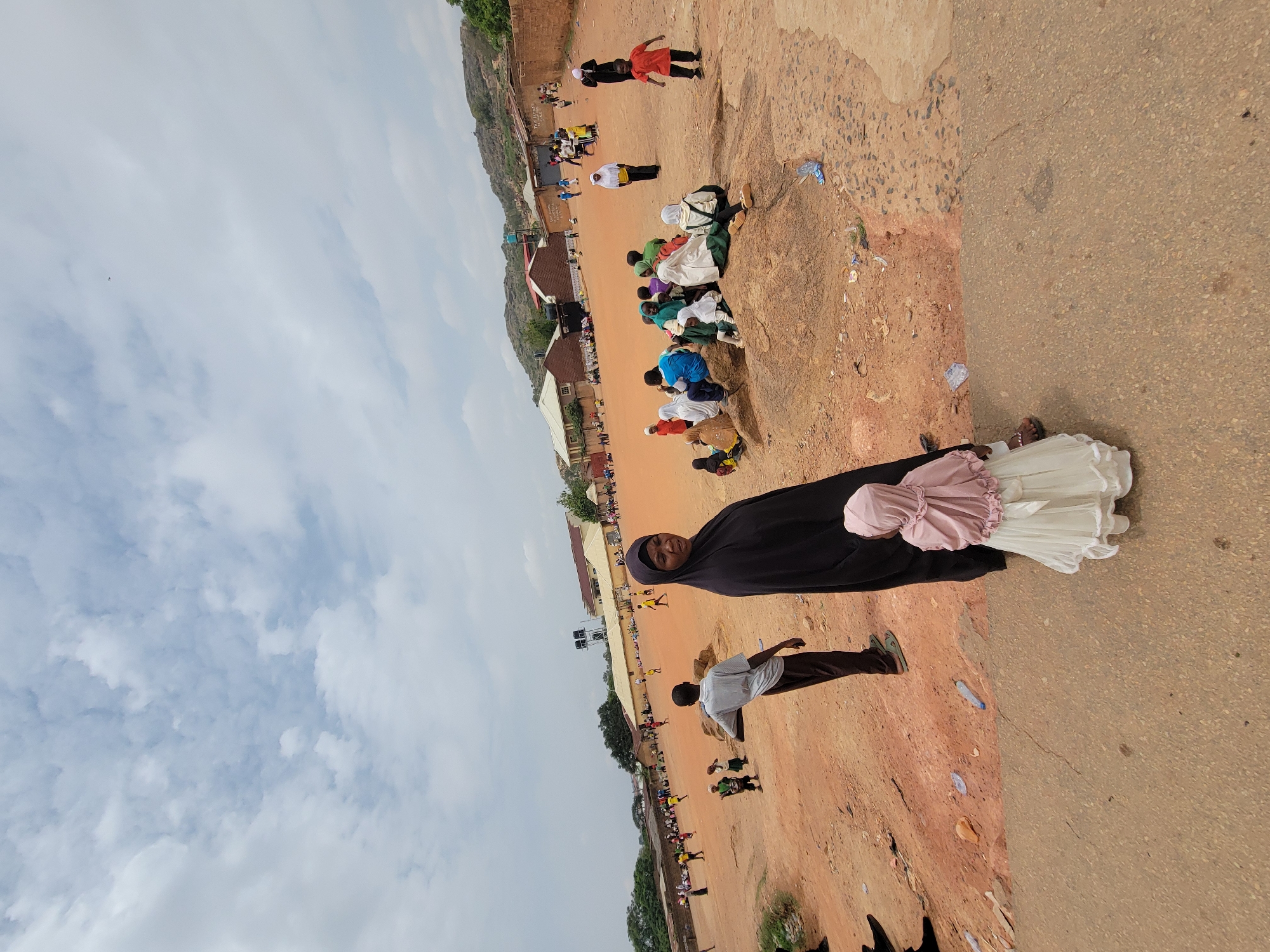 The image shows an outdoor scene, likely in Jos, Nigeria, based on the environment and demographics. A woman wearing a black hijab, a pink dress, and a white garment is in the foreground. A man walks near her, dressed in a white shirt and brown pants. The background reveals a large gathering of people in an open, dusty area. Some individuals are sitting, while others are standing or walking. There are buildings nearby and some vegetation. The sky is overcast. The ground is a mix of dirt and paved road. Several water bottles are scattered on the ground.