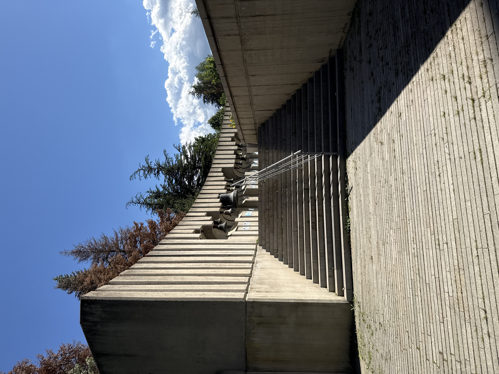 The image shows a concrete structure with a series of vertical fluted elements forming a curved wall. In niches along this wall, several large bells are mounted. A wide set of stone stairs ascends towards these bells, with a metal handrail on the left side. The stairs and the area at their base are paved with horizontal stone slabs, some of which have small plants growing between them. The scene is illuminated by sunlight, casting a distinct shadow across the paved area in the foreground. In the background, there are trees, some appearing dry and brown, against a bright blue sky with scattered white clouds. This is the monument "Bells" located in the city of Bistritsa, Bulgaria. There are no people visible in the image, and no actions or interactions are taking place. The environment is outdoors on a sunny day.