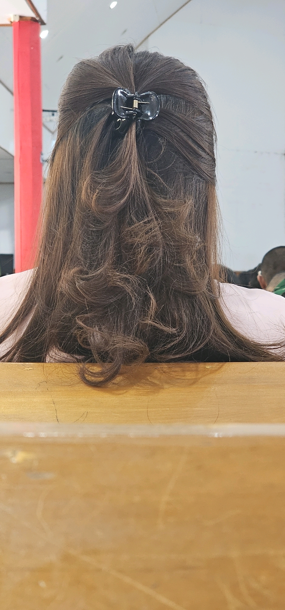 The image shows the back of a person's head with brown, wavy hair held by a black hair clip. The person is wearing a light pink shirt. The hair is styled in loose curls and cascades down. The background includes a blurred red pole on the left and a white wall. In the bottom half of the image, a wooden surface is visible. The context does not suggest a specific location in Puncak Jaya, Indonesia.