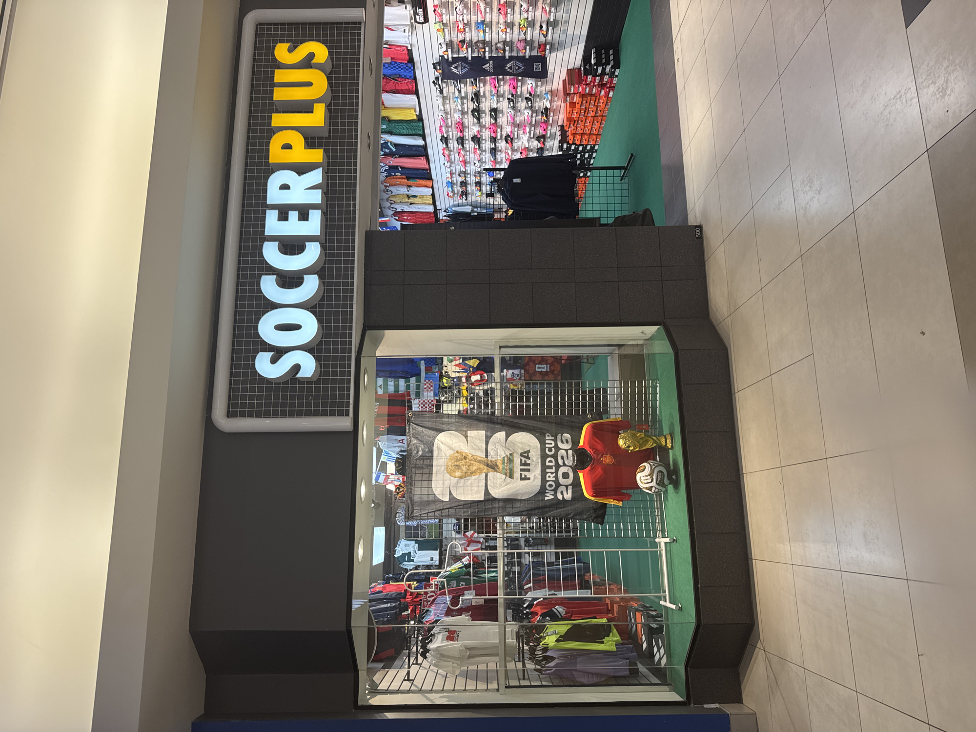 The image shows the exterior of a Soccer Plus store, located in Surrey, Canada. The store features a large display window showcasing soccer apparel and merchandise, including jerseys, a soccer ball, and a trophy. The store's sign, illuminated in blue and yellow, reads "SOCCERPLUS". Inside the store, shelves are stocked with soccer shoes and accessories. The store appears to be located in an indoor shopping mall, as evidenced by the tiled flooring and the overhead lighting. No people are visible in the image.