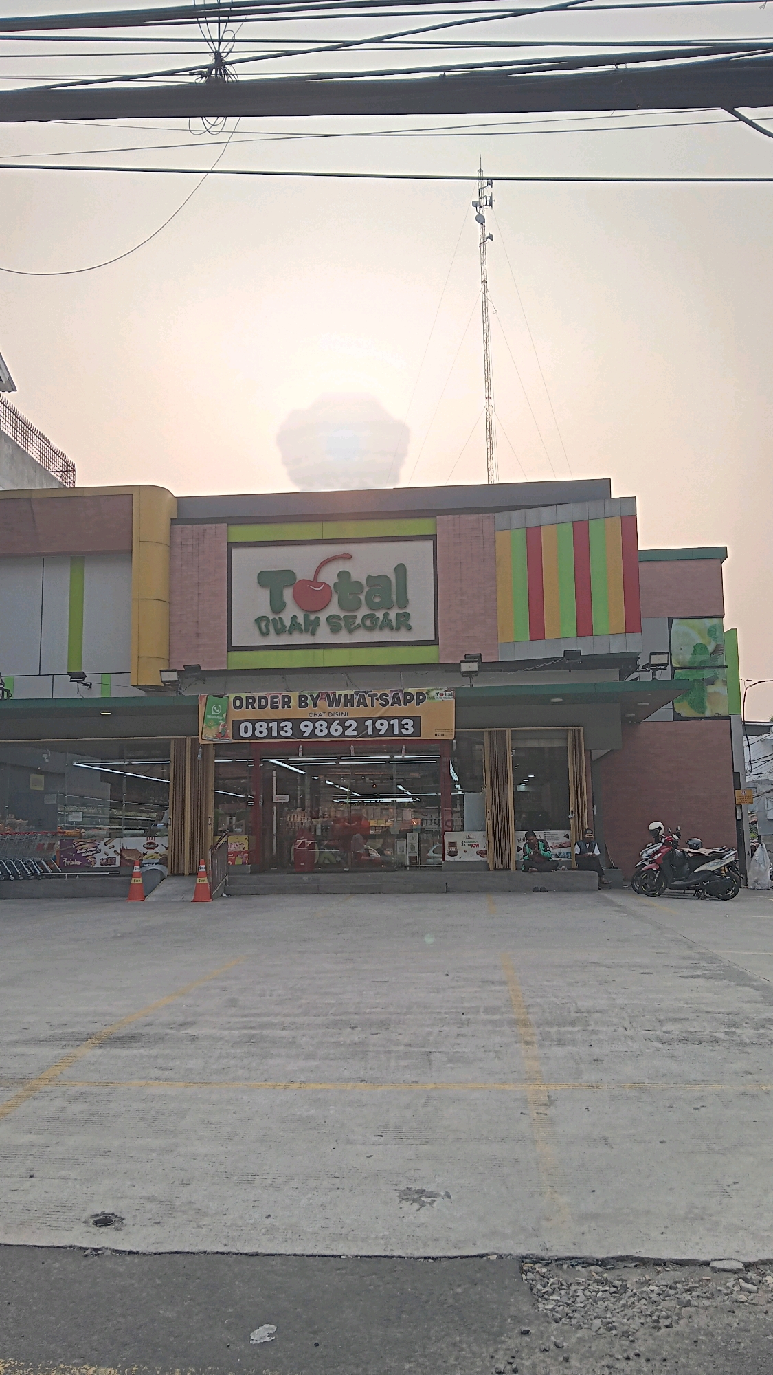In South Jakarta, Indonesia, a frontal view of a "TOTAL BUAH SEGAR" supermarket is captured in the afternoon. The store features a prominent sign with a red cherry graphic and green text. Below the sign, a banner advertises "ORDER BY WHATSAPP" with a phone number, "0813 9862 1913". The store entrance is a glass storefront with visible aisles and displays inside. To the left, a row of shopping carts is parked. Orange traffic cones are placed near the entrance. In front of the store is a paved parking lot with yellow markings. A red motorcycle is parked on the right side of the frame, with two individuals sitting on the curb nearby. The sky is hazy with a large cloud formation obscuring the sun, and power lines and a tall telecommunications tower are visible against the sky.