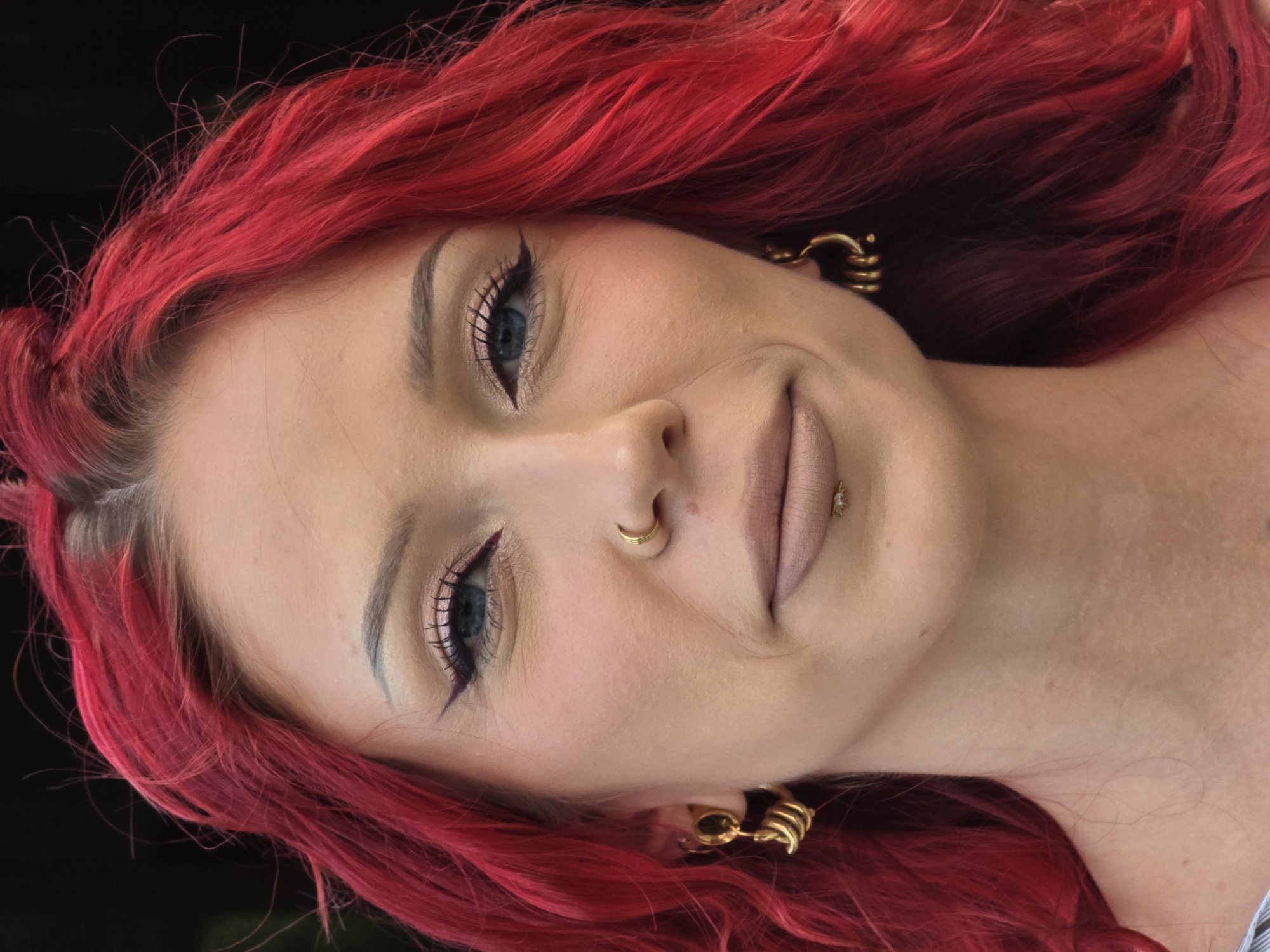 This close-up portrait features a person with vibrant, bright red hair, tilted significantly within the frame against a dark, likely black, background. The individual has fair skin and is looking directly at the viewer with striking blue eyes.
Their face is adorned with prominent makeup, including sharply winged black eyeliner, mascara on long lashes, well-defined eyebrows, and a light, nude-toned lipstick or lip gloss. Several metallic accessories are visible: a gold hoop nose ring in the right nostril, and a small, sparkling stud piercing on the lower lip (labret). Both ears feature distinct gold earrings, which appear to be multi-banded or coiled hoops.
The person displays a subtle, gentle smile. The vivid red hair, which appears to be dyed and styled, creates a strong visual contrast with the dark background. The lighting is bright and even, highlighting the individual's facial features and the intense color of their hair. No text is visible in the image.