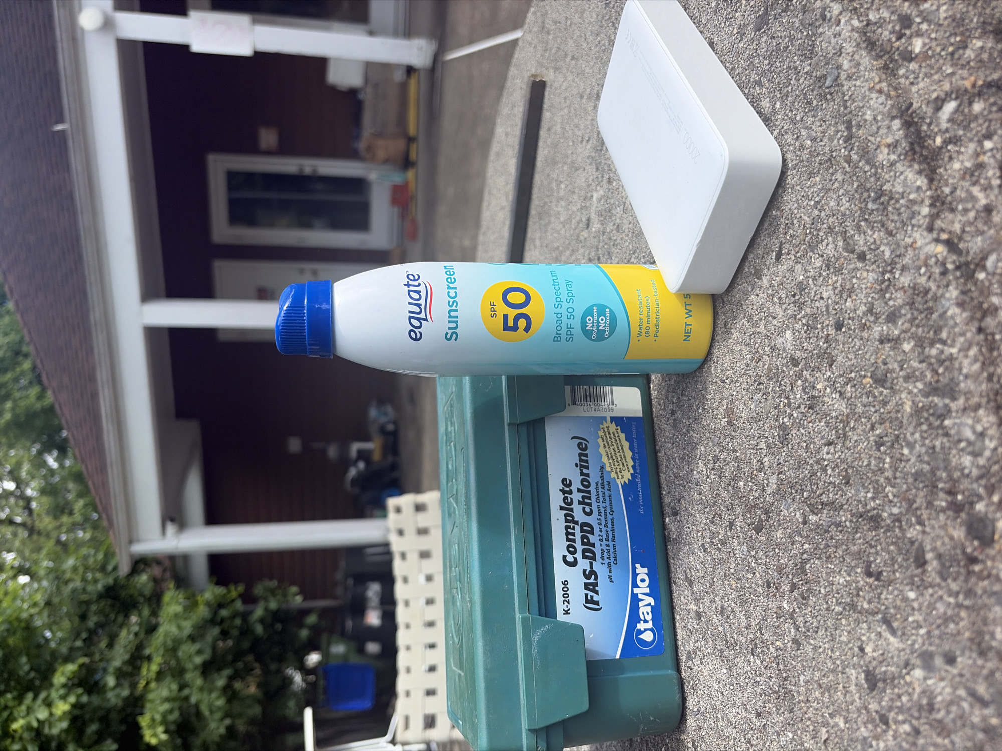 A slightly high-angle, close-up outdoor shot shows an assortment of objects resting on a gray concrete surface. These include a green Taylor K-2006 Complete (FAS-DPD chlorine) water testing kit, a yellow and light blue Equate SPF 50 sunscreen spray bottle, and a white, rectangular electronic device, possibly a portable charger, situated behind the sunscreen bottle. The objects appear to be haphazardly placed, suggesting a temporary arrangement.
In the blurred background, a portion of a two-story building with white support pillars and a dark-colored exterior is visible. Some greenery, possibly trees or shrubs, flanks the left side of the building, suggesting a residential or suburban setting. Other details, such as a ladder and a blue container, are discernible in the background.
While the image offers clues about the setting and objects, a precise location within Edison, United States of America, cannot be definitively determined without additional information or context.
