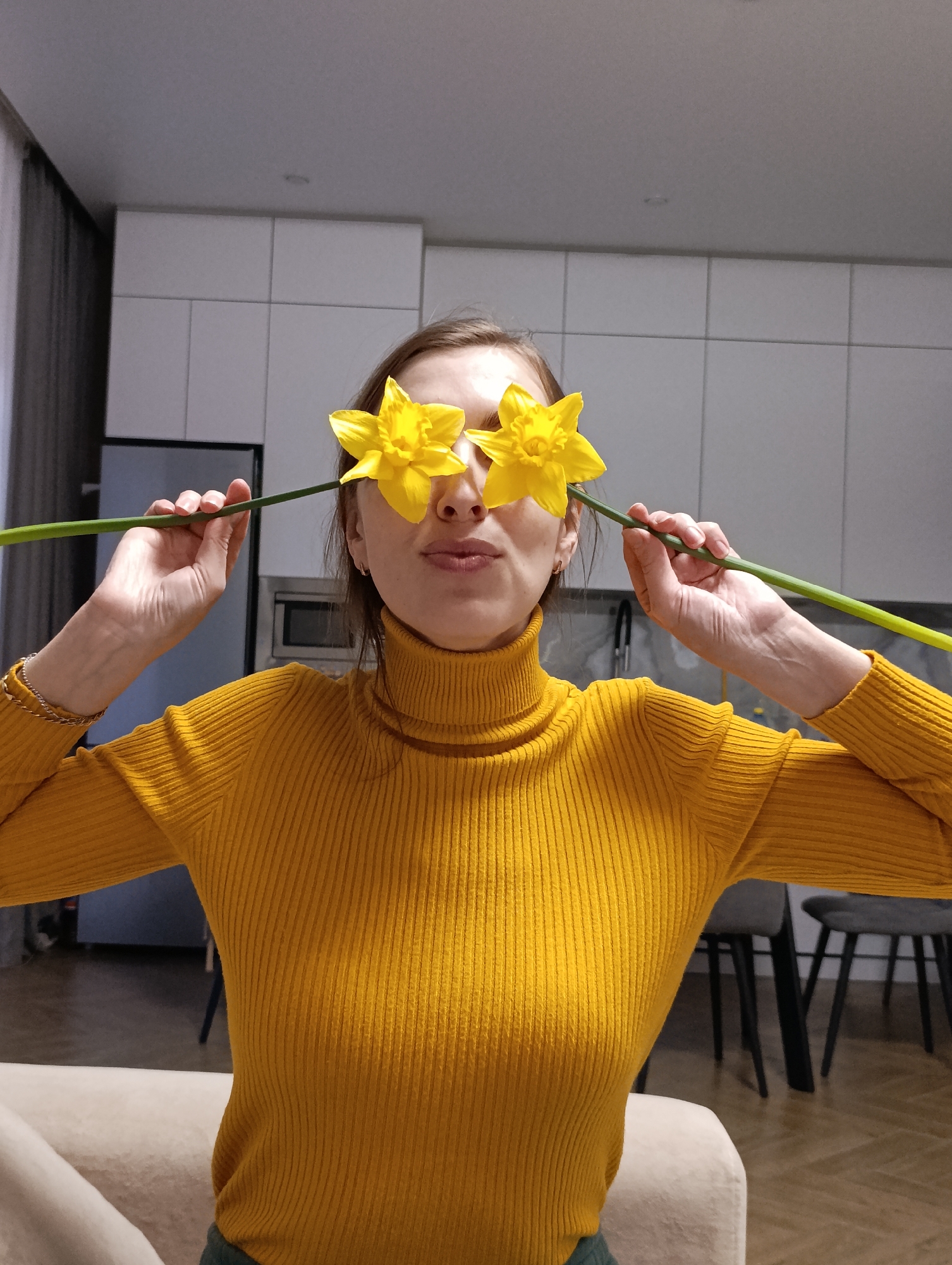 A woman is holding yellow daffodils over her eyes in an indoor setting. She is wearing a yellow turtleneck sweater. The background shows a kitchen area with white cabinets, a stainless steel refrigerator, and a microwave. There is also a dining area with chairs visible. The setting appears to be a home interior. There is no explicit evidence that this image was taken in the city of Chaikovsky, Russia, or any information in the image that would allow to geolocate it.