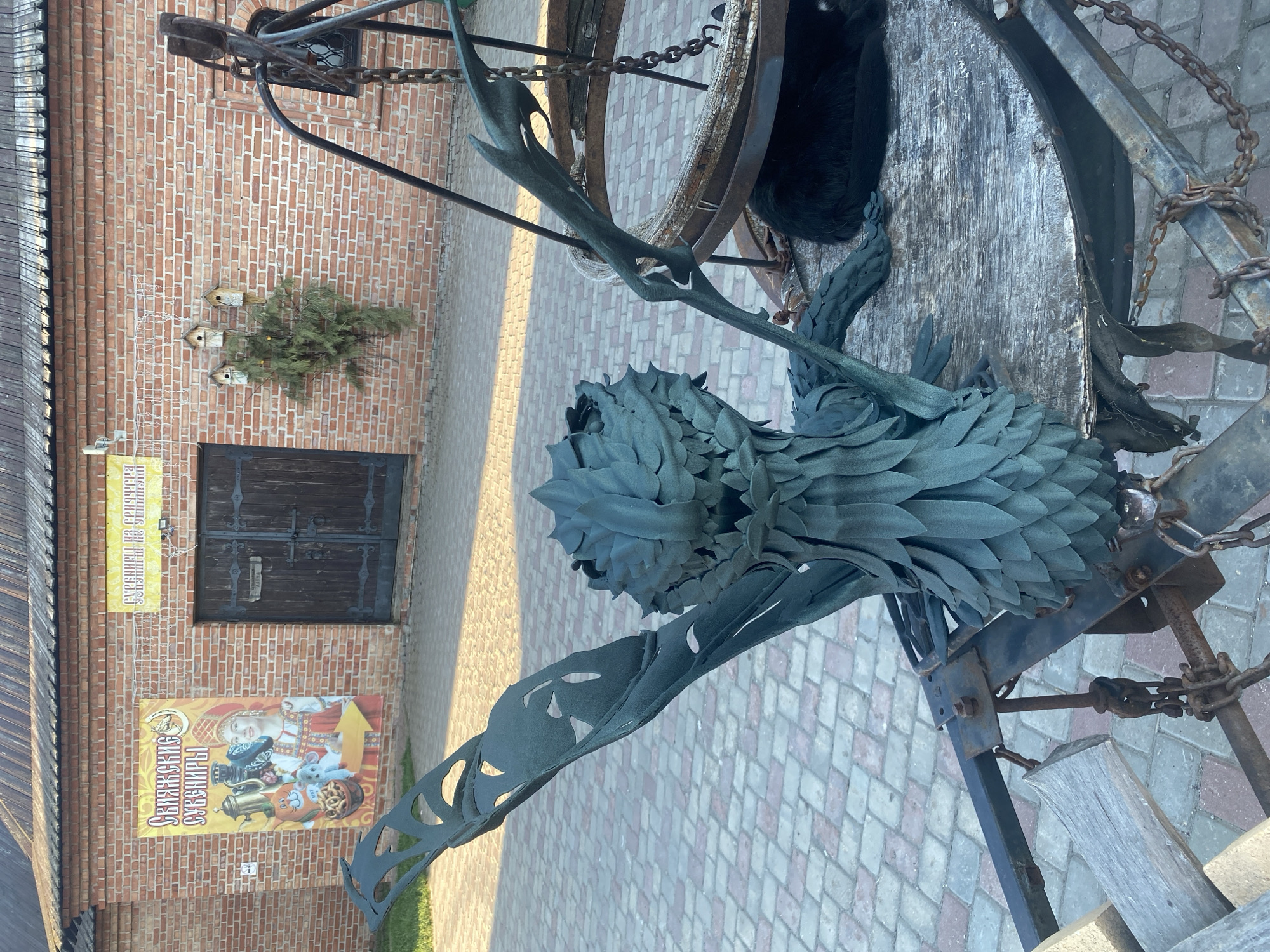 The image shows an outdoor scene in Sviyazhsk, Russia, featuring a stylized metal sculpture of a mythical creature resembling a dragon or bird with feathered or scaled skin, mounted on a rustic metal and wood base. The sculpture is positioned on a brick-paved surface. Behind the sculpture, a brick building is visible, adorned with a sign that includes Cyrillic text and images, likely indicating a local souvenir shop or attraction. There are also birdhouses mounted on the wall, suggesting a natural or decorative theme. The scene is bathed in sunlight, casting shadows across the paving stones. In the background, part of an iron wheel with chained components is visible. A dark patch, possibly fur or fabric, is lying on top of the wheel. The overall impression is that of a cultural or tourist location, with unique artistic elements within a historical setting.