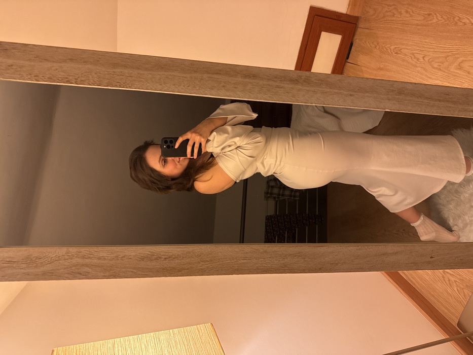 A female individual is captured in a mirror selfie within an indoor setting in Cava de' Tirreni, Italy. She wears a light-colored, off-the-shoulder dress with long sleeves, form-fitting around the torso and hips, flaring near the calves, and white frilly socks. A black bra strap is visible on her left shoulder. The subject holds a dark smartphone with both hands, aiming it towards the mirror, with a slight smile directed at the phone's screen. The mirror features a light wooden frame. Reflected within the mirror, behind the individual, is a warm-toned lamp with a rectangular fabric shade, dark patterned clothing folded on a surface, and a light-colored bedspread. A white shaggy rug is underfoot, with a wooden floor visible beyond. A brown rectangular object, likely a frame, lies on the wooden floor to the right of the reflected scene. The room's ambient lighting is warm, and the wall opposite the mirror is a light pinkish-beige with a wooden baseboard.