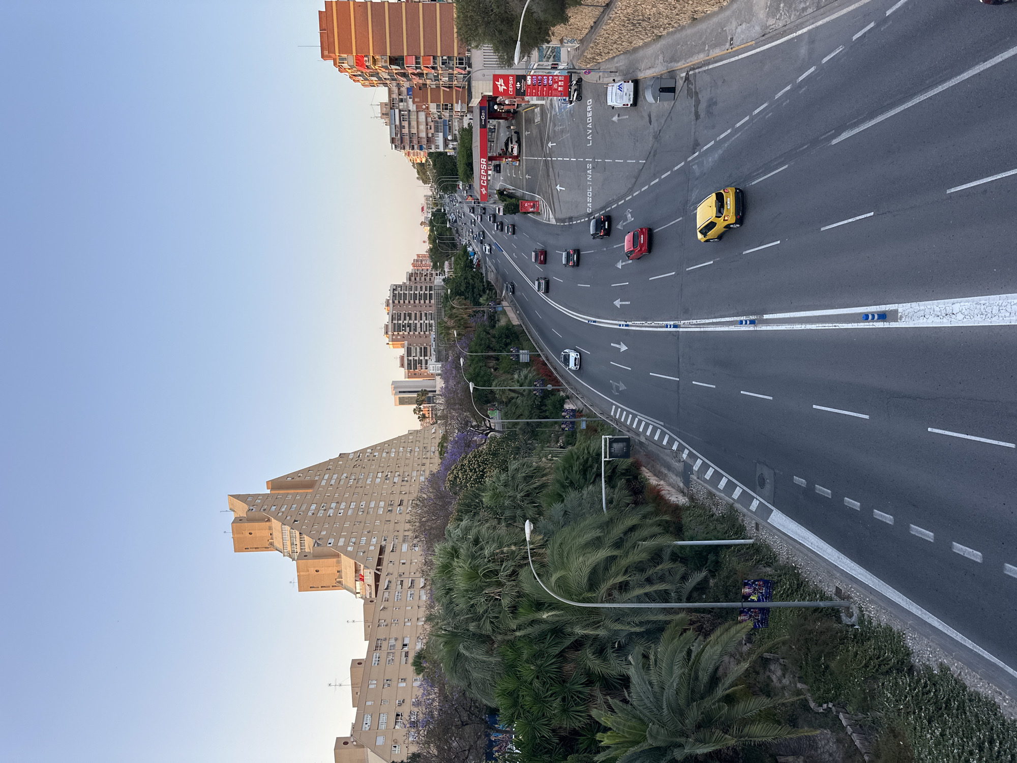 An aerial view captures a multi-lane road in an urban setting under a clear blue sky, likely during the late afternoon or early evening based on the lighting. The road, paved in asphalt, is marked with white lines indicating lanes and direction. Several vehicles, including cars and a white van, are present on the road, some in motion and others parked.
To the left of the road, lush green palm trees and other vegetation are visible, bordering a walkway and a stone wall. Beyond the greenery, a distinctive, angular, and stepped building with numerous windows dominates the landscape, its beige facade reflecting the ambient light. This building is complemented by other mid-rise apartment buildings with balconies and window arrays. In the distance, more urban structures and a gas station with red and white signage are discernible.
On the right side of the road, a row of brick buildings with orange awnings lines the street. A stone wall with vegetation at its base separates the road from these buildings. The overall scene depicts a bustling urban environment with a clear division between the road, natural elements, and residential or commercial structures.
