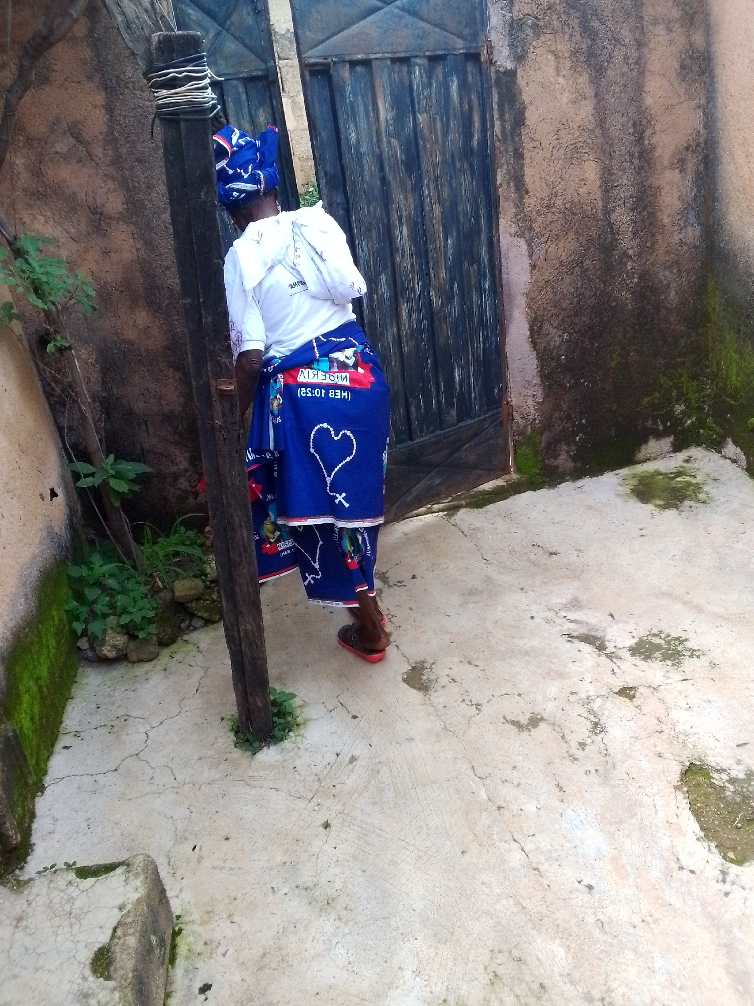 A woman wearing a white t-shirt and a blue patterned skirt with white writing and a heart design is standing next to a wooden pole in an outdoor setting. She is facing away from the camera, looking towards a dark, vertical-slatted gate. The ground is made of cracked concrete with some moss and green vegetation growing in patches. A stone wall, partially covered in moss, runs along the left side of the frame, and another wall of similar texture is visible to the right, behind the gate. The overall scene appears to be a back alley or courtyard.