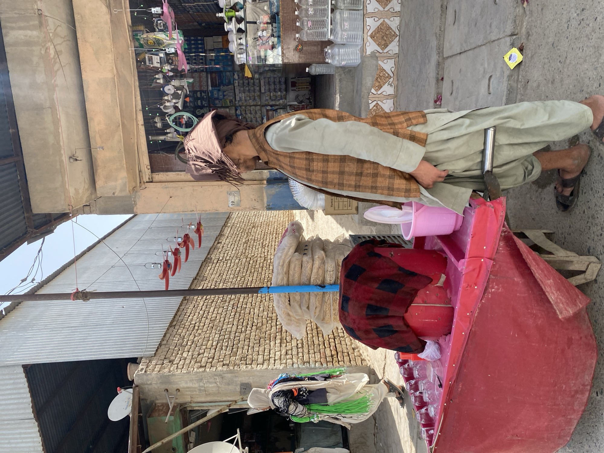 The image depicts a street scene in the city of Kandahar, Afghanistan. In the foreground, a man wearing a light green tunic, a brown checkered vest, and a brown headscarf stands next to a bright red street cart. He has a knife tucked into the waistband of his pants. The cart has glasses and a pink bucket on it. Behind him, a shop entrance is visible, displaying various goods, including lights and water bottles. To the left of the cart, another person, dressed in traditional Afghan clothing, walks away from the camera, carrying bundles of green material and wires. In the background, a building with a corrugated metal roof and stone walls is visible, along with satellite dishes. The ground is paved and the sky is clear.