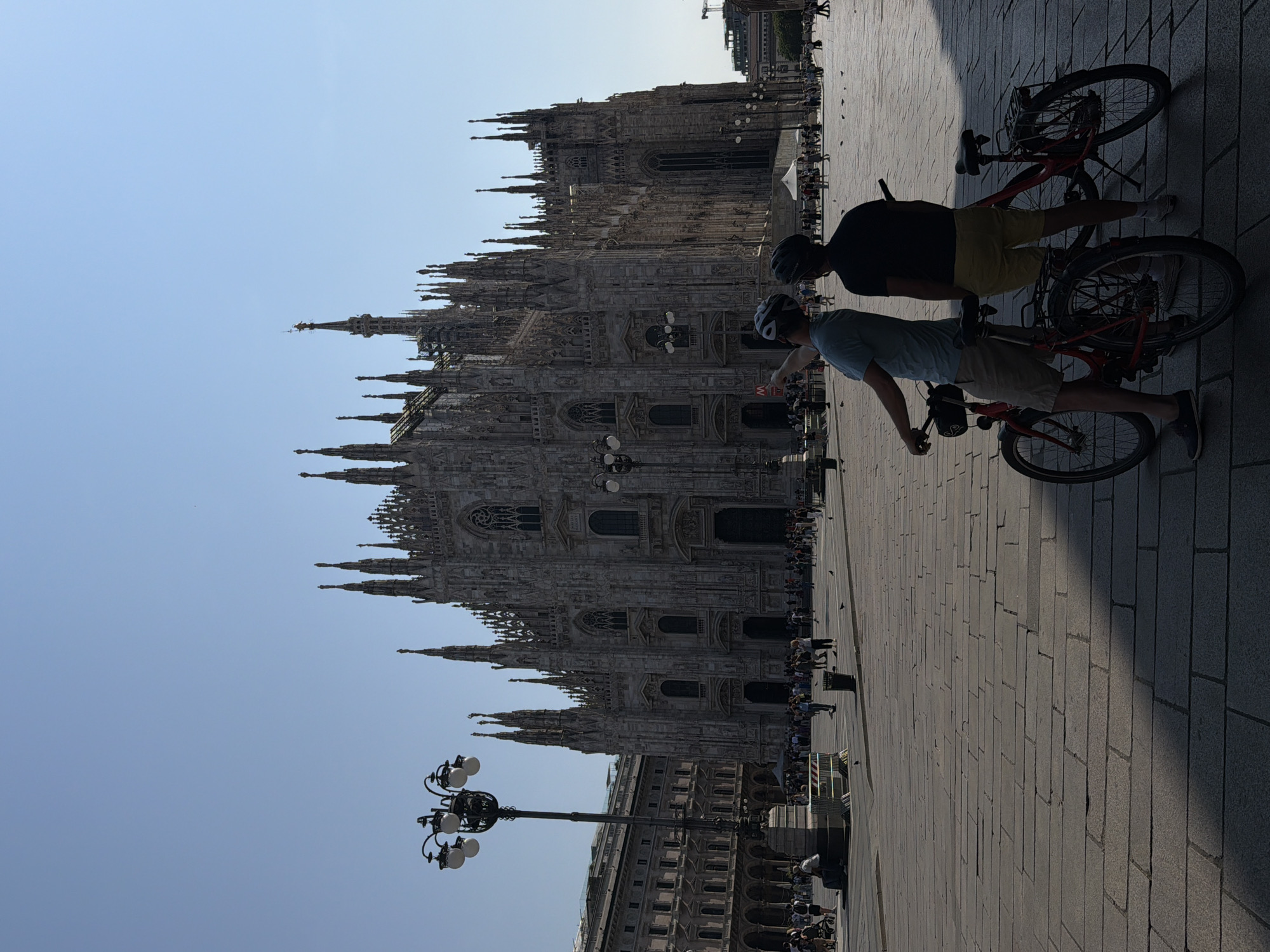 Two people on bicycles are in the foreground of the image. They are wearing helmets and casual clothing. Behind them, towering high, is the Duomo di Milano, an enormous cathedral. A lamppost is also visible closer to the cathedral. People walk along the perimeter of the building. The pavement is made of large blocks. The sky is clear and light blue.