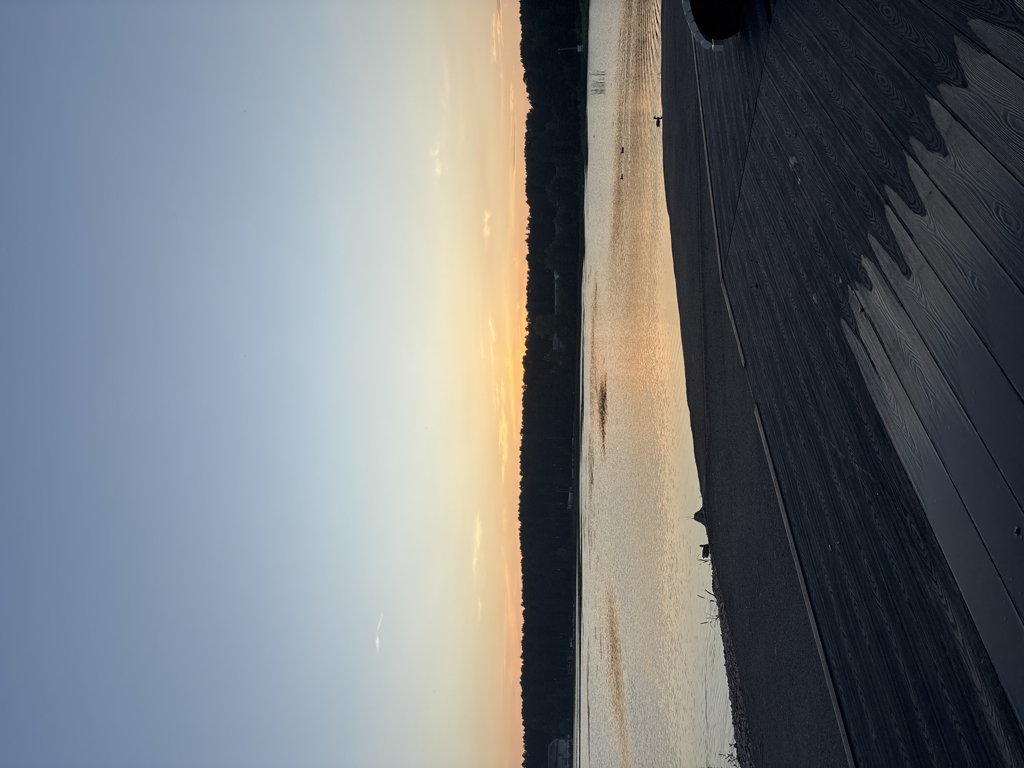 The image depicts a tranquil lakeside scene at dusk in Zheleznogorsk, Russia. The sky is a gradient of soft blues and oranges, transitioning from a clear blue zenith to a warm, diffused sunset glow near the horizon. A dark line of trees borders the far side of the calm lake, their silhouettes stark against the illuminated sky. In the foreground, a dark wooden deck with visible wood grain stretches across the bottom half of the frame, angled from right to left, suggesting a waterfront viewpoint. The surface of the deck appears slightly damp in patches, reflecting the subdued light. The water itself is a muted silver, rippling gently and catching the colors of the sunset. Two ducks are visible on the water, small dark shapes in the vastness of the lake, and a third duck stands on the gravelly shore at the edge of the water. The overall impression is one of quietude and natural beauty.