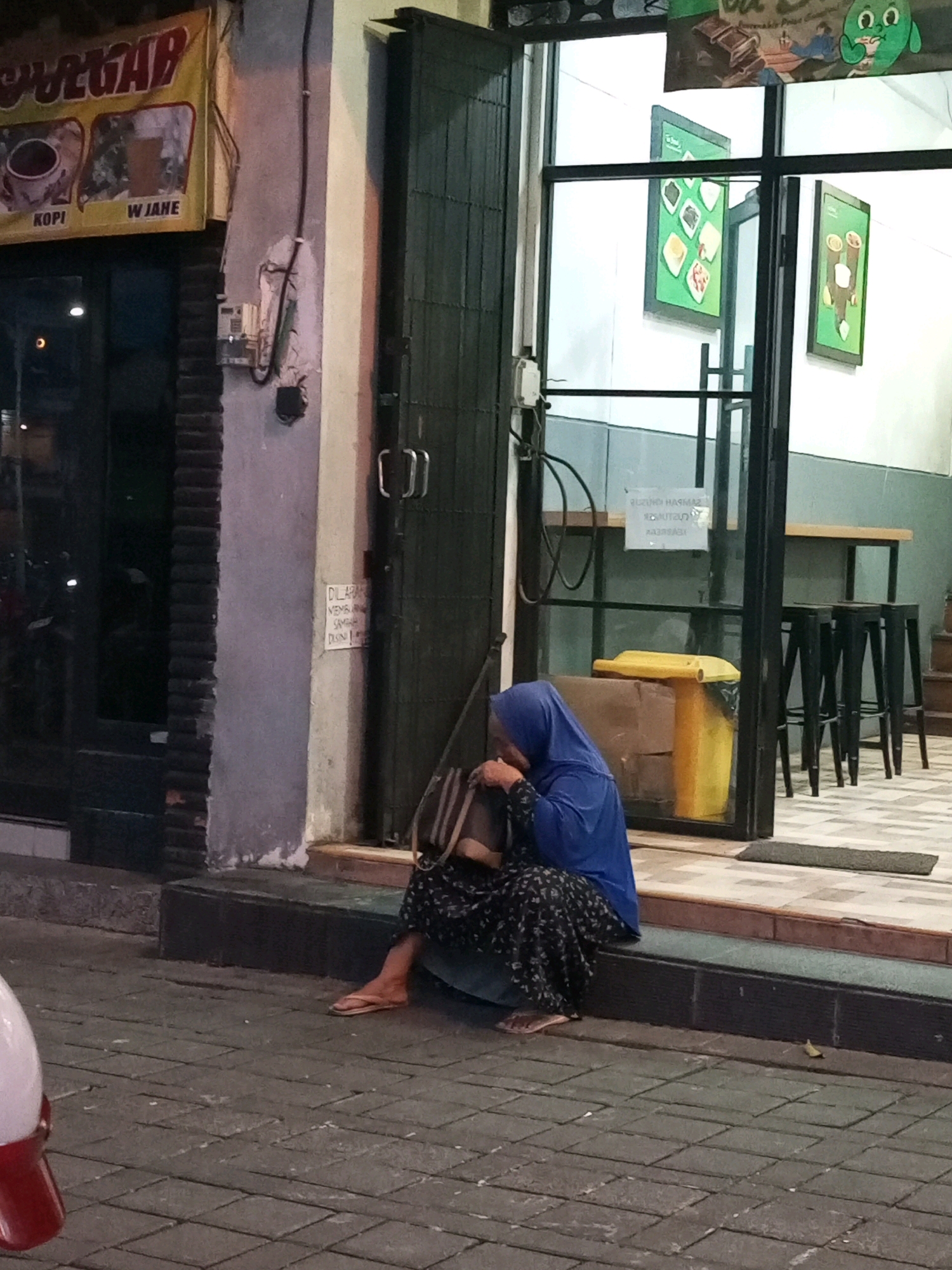 The image shows a woman wearing a blue hijab and a patterned skirt, sitting on a raised concrete step outside a shop. She is holding a bag and appears to be resting. In the background, the shop has large glass doors and windows, revealing the interior with tables, chairs, and framed pictures on the wall. A yellow trash can and cardboard boxes are visible near the entrance. A sign with Indonesian text, "DILARANG MEMBUANG SAMPAH DISINI," meaning "DO NOT DUMP TRASH HERE," is posted on the wall to the right of the entrance. The shop appears to be located on a street with a cobblestone pavement. The context suggests this is a street scene in Batu, Indonesia, given the typical architecture and signage found in such locations.