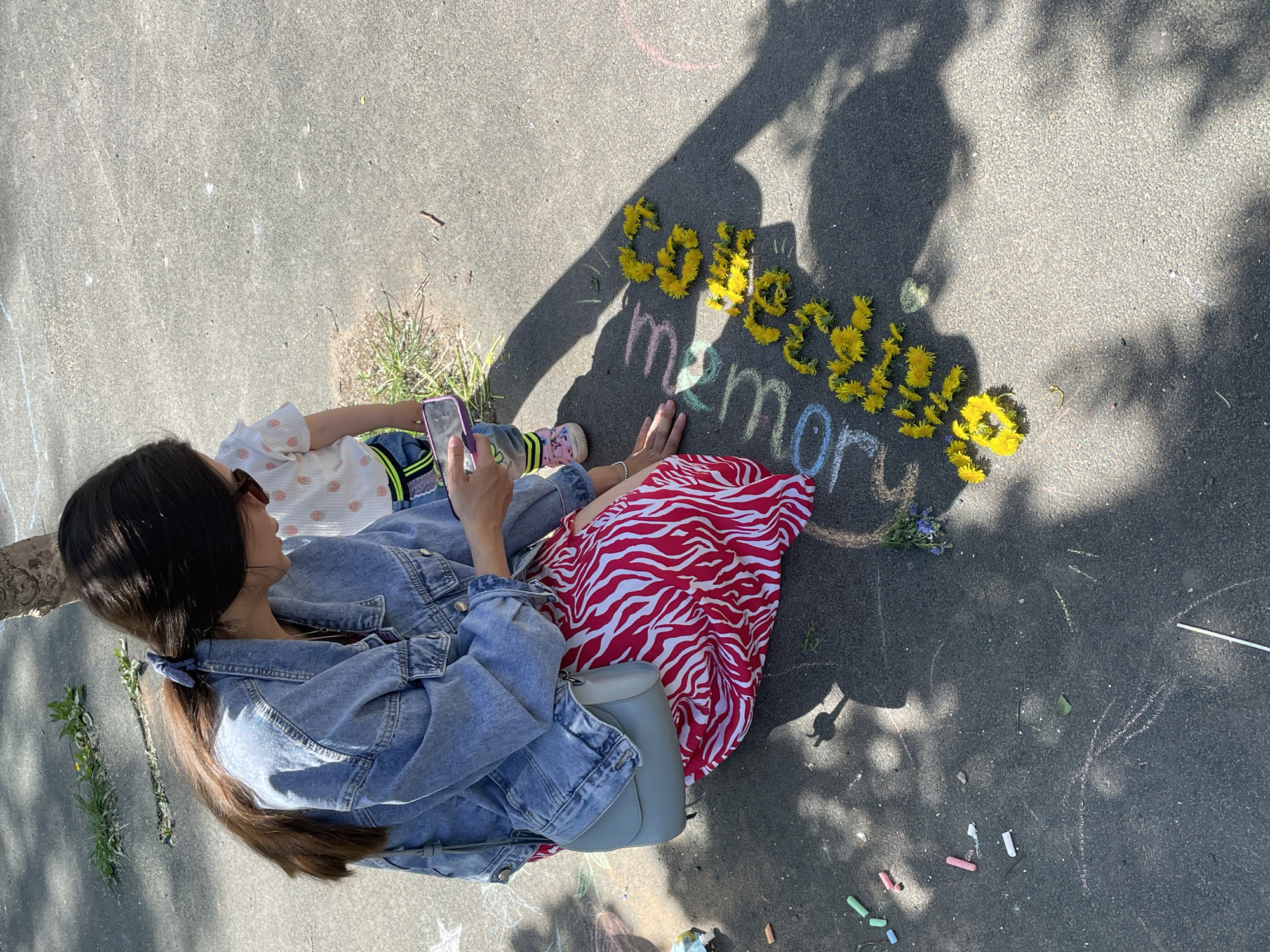 A high-angle, full shot depicts a woman and child sitting on asphalt next to the chalked and floral text "Soviet memory." The woman, seated with her legs extended, wears a denim jacket, a red and white zebra-striped dress, and has a blue purse beside her. She is holding a smartphone in her hands and appears to be taking a photo. A child, wearing a light-colored top with a strawberry print, jeans, and pink shoes, sits next to her. The text "Soviet" is spelled out in yellow dandelions, while "memory" is written in colored chalk below. Several sticks of colored chalk are scattered on the asphalt near the text. A shadow obscures part of the text and surrounding asphalt. The location in Izhevsk, Russia, is not verifiable from the image.