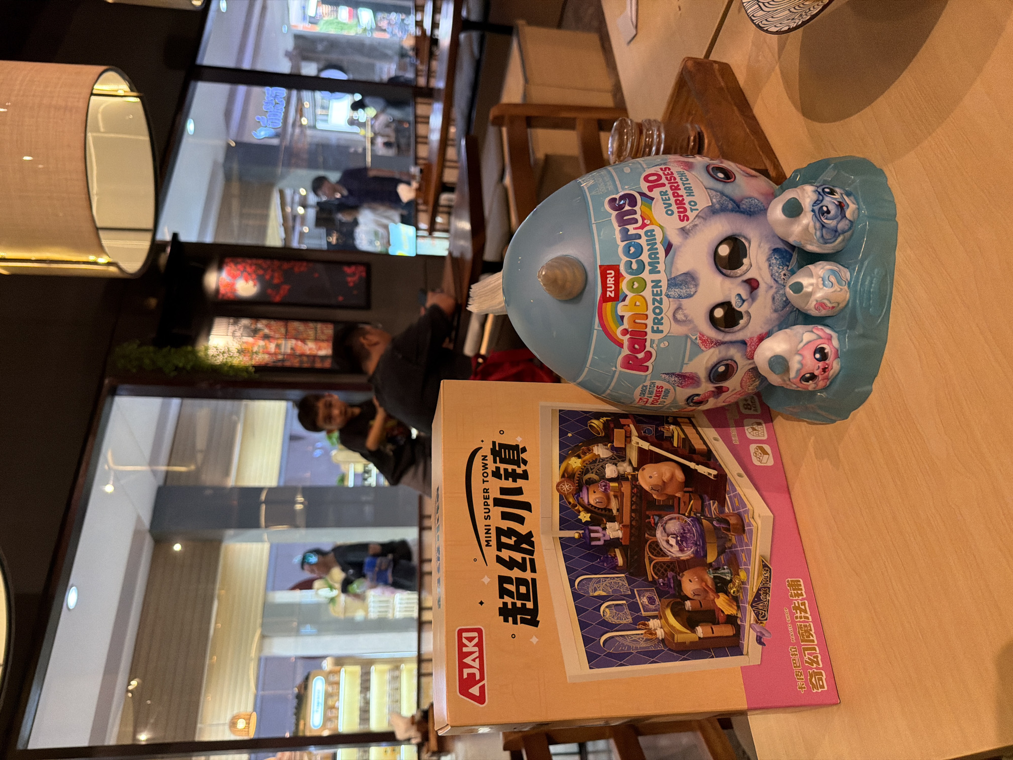 In a bright, well-lit indoor establishment, possibly a restaurant, as suggested by the wooden tables and chairs, two children are partially visible in the background, engaging in an activity. On the foreground table, a vibrant orange box of "JAKI MINI SUPER TOWN" building blocks, featuring Chinese characters, is positioned next to a large, blue, egg-shaped toy. The toy is identified as "ZURU Rainbocorns FROZEN MANIA," boasting "OVER 10 SURPRISES TO HATCH!" The scene is viewed from a medium-high angle, with a shallow depth of field that blurs the background and focuses attention on the toys. The overall impression is one of a retail or play environment, possibly a souvenir shop or a toy section within a larger establishment. The specific location of Kuta, Indonesia, is not visually discernible from the image itself.