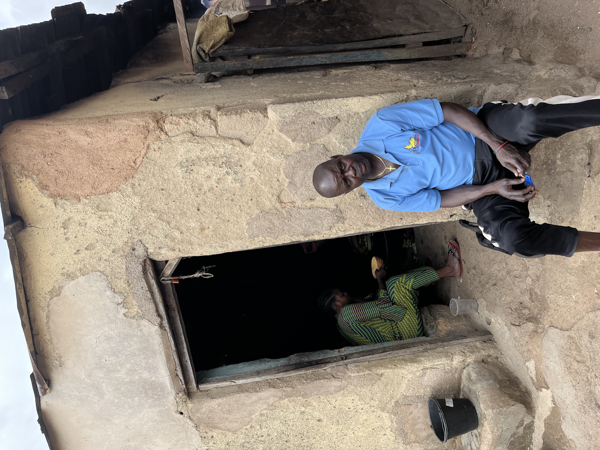 The image, originally rotated 90 degrees clockwise, captures a daytime scene in Jos, Nigeria, showing an adult male seated outside a building and another person partially visible inside a low window.
In the foreground, an adult male with a bald head is seated against the rough-textured wall of a building. He wears a light blue short-sleeved polo shirt, black athletic trousers with a white stripe down the side, and a gold chain with a cross pendant. A red bracelet adorns his left wrist. He looks directly at the camera with a neutral expression, holding a small blue object in his hands, possibly a phone or a small container. The logo on his shirt clearly reads "Operation Rainbow," accompanied by a graphic of a bird or dove.
The building's exterior wall is constructed of rough concrete or stone, showing various patches of light yellow, light brown, and pinkish plaster. Below the man, near the base of the wall, a small black bucket and a clear plastic bottle are visible on the ground.
To the man's right, a dark, rectangular window opening reveals a second individual inside. This person, partially visible, is wearing a vibrant green, black, and orange patterned garment. They appear to be seated on the floor and are holding a light-colored, round object, possibly food or a bowl. The interior of the room beyond the window is dimly lit.
Above the wall, a simple, rustic roof structure made of dark wooden beams and possibly corrugated metal is visible. A light-colored sack with some faint, unreadable text is draped over a portion of the roof. The lighting suggests it is daytime, with diffused light indicating either an overcast sky or bright ambient conditions, as there are no strong shadows. The overall setting depicts a modest, possibly residential structure in an urban or semi-urban area of Jos, Nigeria.