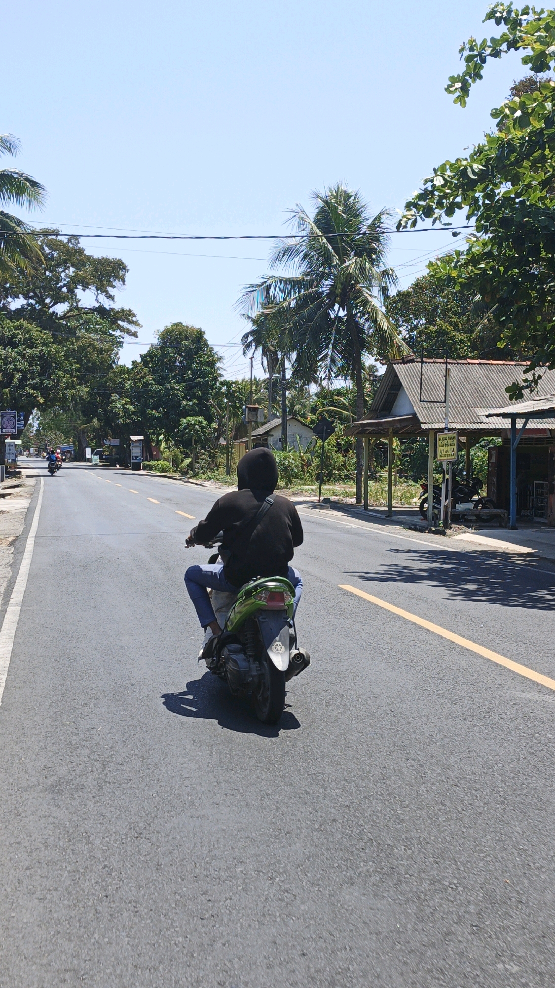 A person wearing a black hoodie and blue jeans rides a green scooter on a paved road in Desa Wonoharjo, Indonesia. The scooter is in the foreground, with the rider's back to the camera. The road is lined with trees and a few buildings, including a structure with a corrugated metal roof. The sky is clear and blue, and the sun is casting shadows on the road. There are other scooters and vehicles visible in the distance. The environment appears to be a rural or semi-rural area with lush vegetation and a tropical climate.