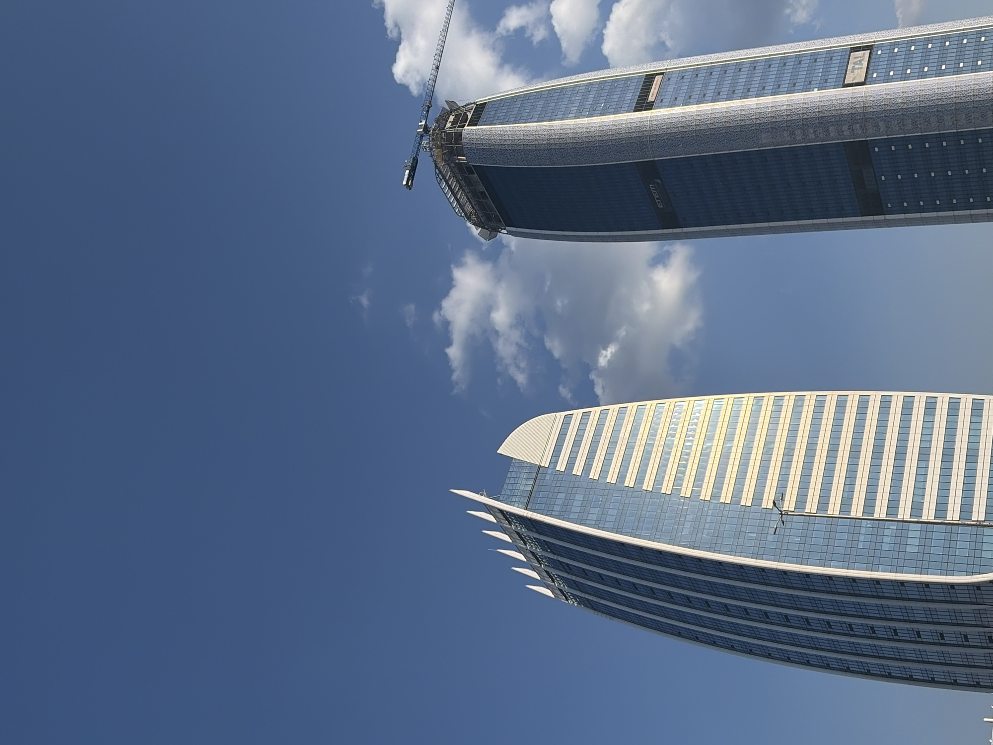This is a media file featuring two modern skyscrapers against a bright blue sky with scattered white clouds. The image appears to be taken from a low angle, looking upwards at the structures.
The top skyscraper is partially obscured by a construction crane, indicating that it is either under construction or undergoing renovation. It has a curved, sleek design with a facade composed of glass panels and metallic accents. The word "emem" is visible vertically on the blue section of the building.
The bottom skyscraper is also modern and features a curved, elongated shape. Its facade is a mix of blue glass and a lighter-colored material with vertical stripes, possibly glass or metallic elements, which reflect the sunlight. A horizontal pole with some fixtures extends from the middle section of this building.
The overall scene suggests an urban environment with significant architectural development. The bright blue sky and sunlight indicate it is daytime, likely a clear or partly cloudy day. There are no people visible in the image.