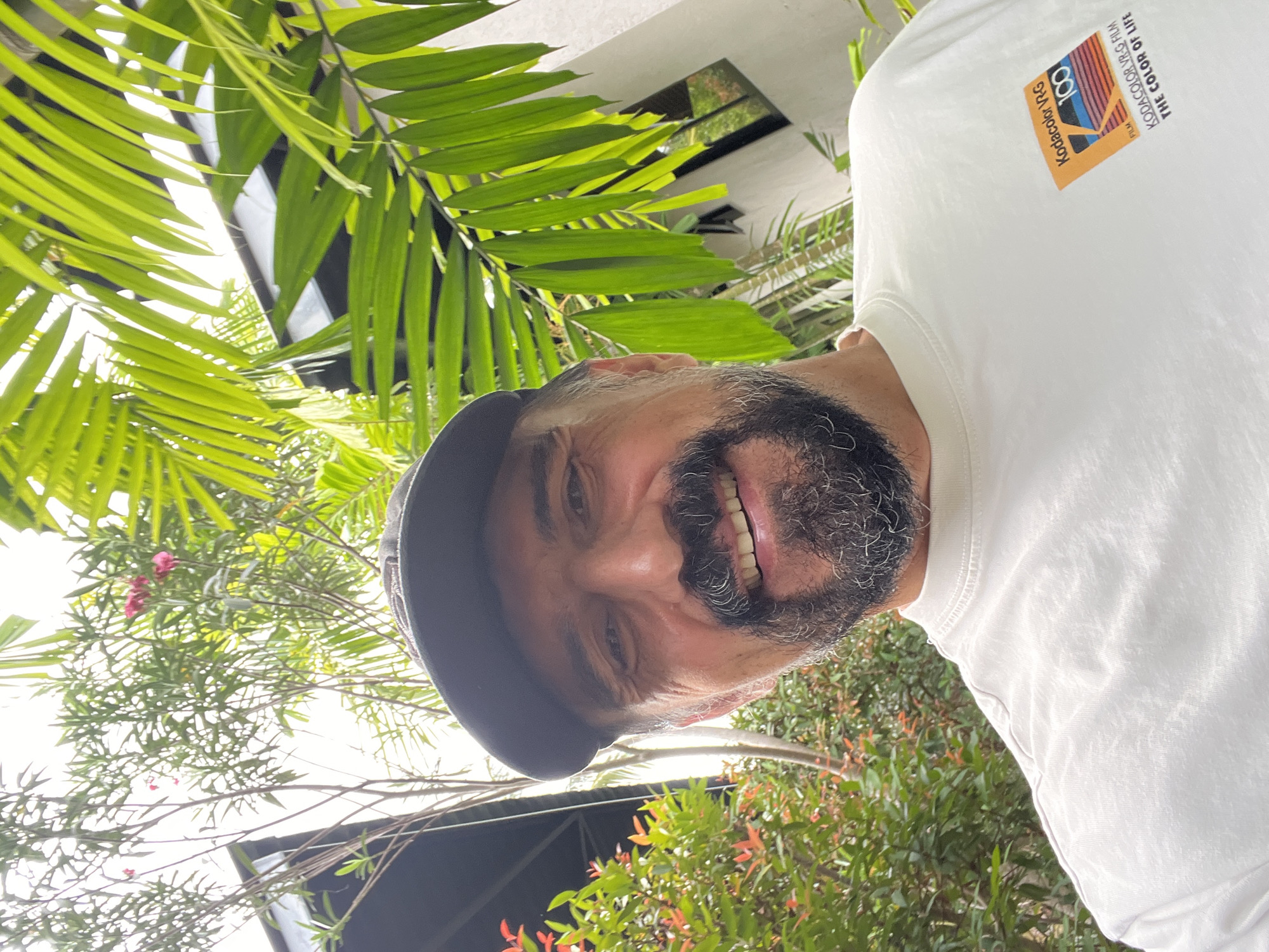 The image features an adult man smiling broadly, captured in a vibrant outdoor setting in บ้านบางรักษ์, Thailand. He has a dark and grey beard, wears a dark cap, and a white t-shirt. His joyful expression is directed towards the camera, suggesting a selfie or a close-up portrait.
The background is characterized by lush tropical foliage, including prominent green palm fronds and other dense leafy plants, with hints of small red or pink flowers. A bright sky is visible through the canopy, indicating it is daytime with abundant natural light and sunny weather. Part of a white building structure can also be seen through the leaves.
A distinctive detail is the graphic on the man's white t-shirt. The visible text, as per OCR, reads: "Kodacolor VR-G 100 FILM KODACOLOR VR-G FILM THE COLOR OF LIFE," referencing the classic Kodak film brand and slogan, adding a nostalgic touch to the image. The overall mood conveyed is cheerful and relaxed.