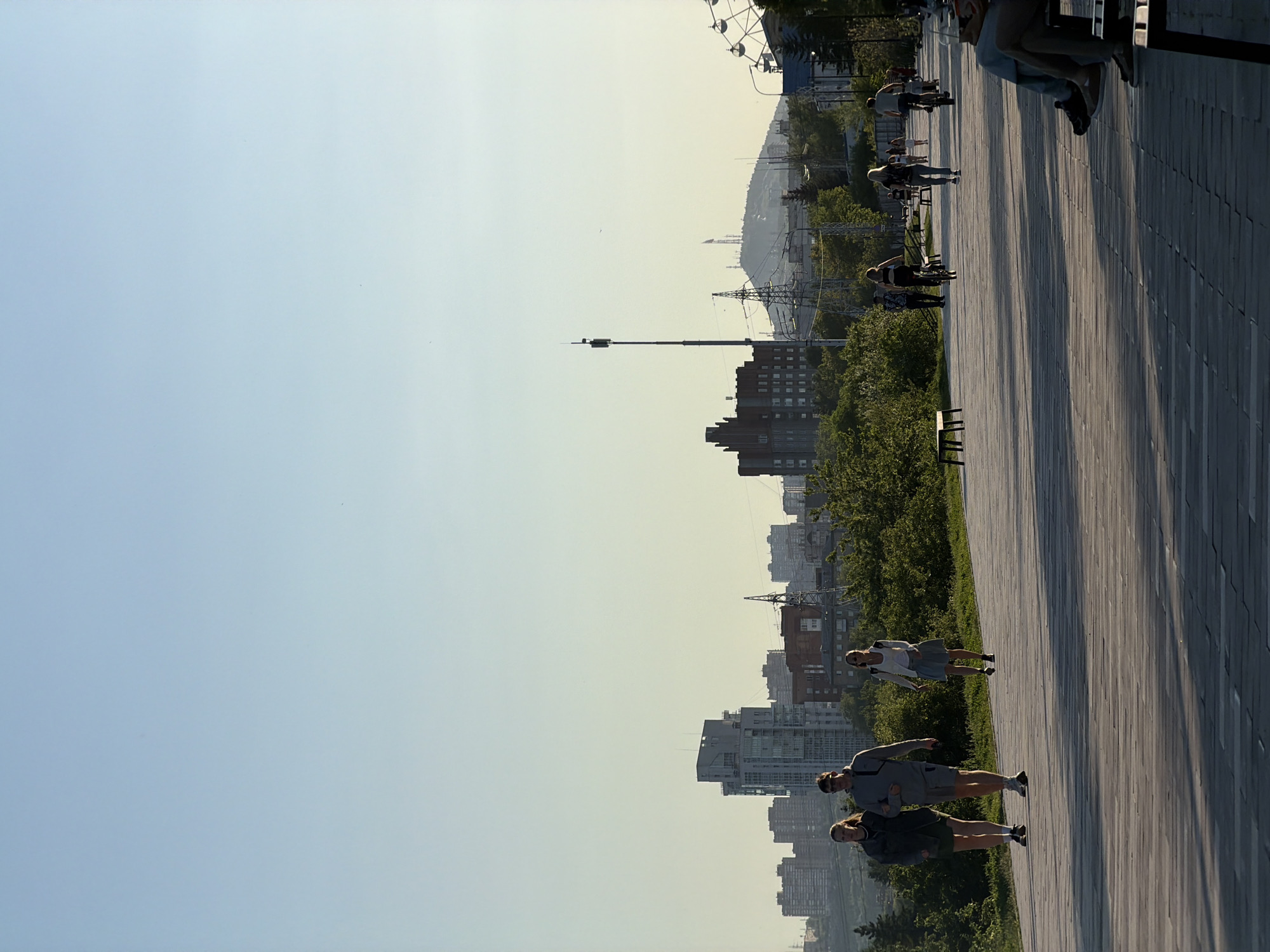 The image depicts an outdoor scene in Krasnoyarsk, Russia, likely an urban promenade or park area, captured during either early morning or late afternoon, as indicated by the long shadows stretching across the ground.
In the foreground and mid-ground, a wide wooden plank path dominates the lower right portion of the frame, extending into the distance. Several people are present on this path: in the immediate foreground, three individuals (two women and one man) are standing. Further along, a woman in a white top and light skirt is walking, and other figures are seen walking, cycling, or sitting on benches in the distance.
To the left of the path, a band of green trees and bushes provides a natural boundary. Beyond this greenery, an urban landscape unfolds with numerous multi-story buildings comprising a city skyline under a hazy sky. A tall, slender communication tower stands prominently among the buildings. In the far background, hazy hills or mountains are visible, defining the horizon. A large Ferris wheel is partially visible on the far right, suggesting a recreational area. Power lines and transmission towers are also noticeable within the urban sprawl.
The sky is clear but appears slightly hazy towards the horizon, contributing to a soft, warm light that illuminates the scene. People on the path appear to be leisurely enjoying the outdoor environment. No discernible text is visible.