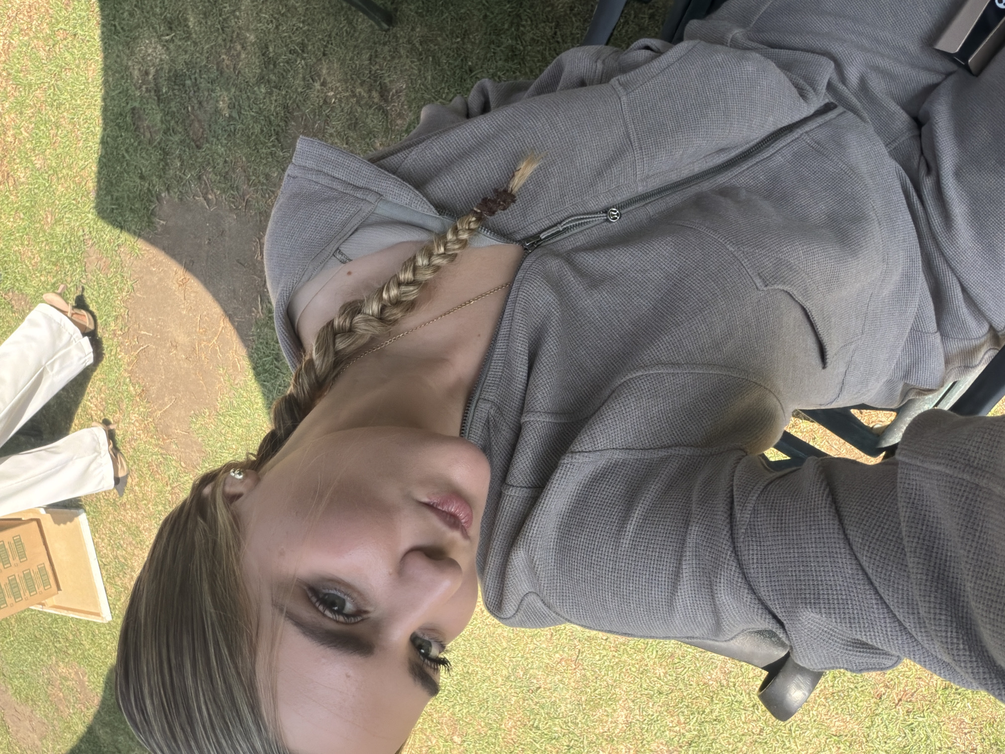 A fair-skinned woman with blonde hair braided in a single plait sits outdoors on a black plastic chair, wearing a grey thermal hoodie and matching sweatpants. A thin gold chain is visible around her neck. She is angled towards the camera, looking directly at the viewer with a slight pout.
To the left of the frame, partially obscured, are portions of white pants and a cardboard box with product labeling. The ground consists of grass and bare earth.
The location is likely a yard or grassy area in San Andrés Cholula, Mexico, based on the information provided, but there are no discernible landmarks in the photograph to confirm this.
