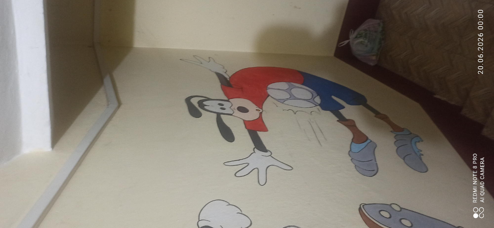 The image depicts a wall decorated with a cartoon mural of Goofy playing soccer. Goofy is depicted in mid-action, kicking a soccer ball. The mural is on a light-yellow wall, and the bottom of the wall transitions to a dark red trim. In the lower right corner of the image, a plastic bag filled with items is placed on a floor with a parquet pattern. The scene is lit by ambient lighting, and a timestamp of "20.06.2026 00:00" is visible in the bottom right corner, along with camera information "REDMI NOTE 8 PRO AI QUAD CAMERA". The room appears to be indoors, possibly a child's room or play area, given the cartoon mural.
