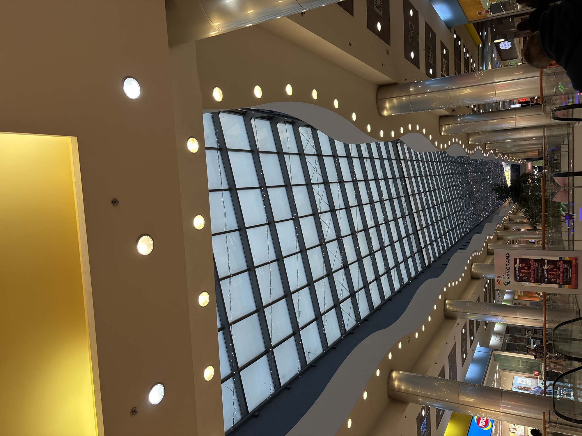 The image shows the interior of the Panorama Mall in Pleven, Bulgaria. A long, skylight-style ceiling structure with a grid pattern runs down the center of the mall. Rows of recessed lights line the edges of the ceiling feature. Storefronts and columns are visible along the sides of the mall. Escalators and informational signs are also visible.